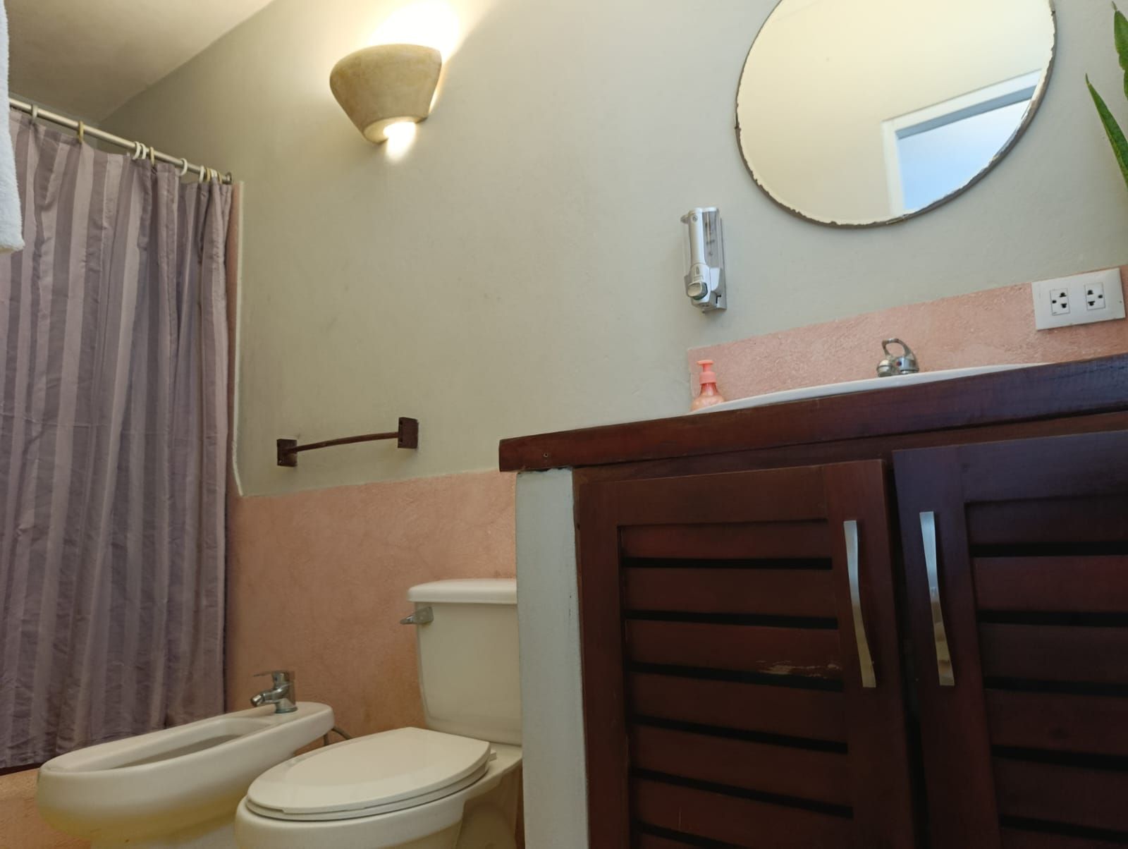 Bathroom with toilet, bidet, sink, mirror, and a wood cabinet. Soft pink walls, tan tile floor, and a pink shower curtain.