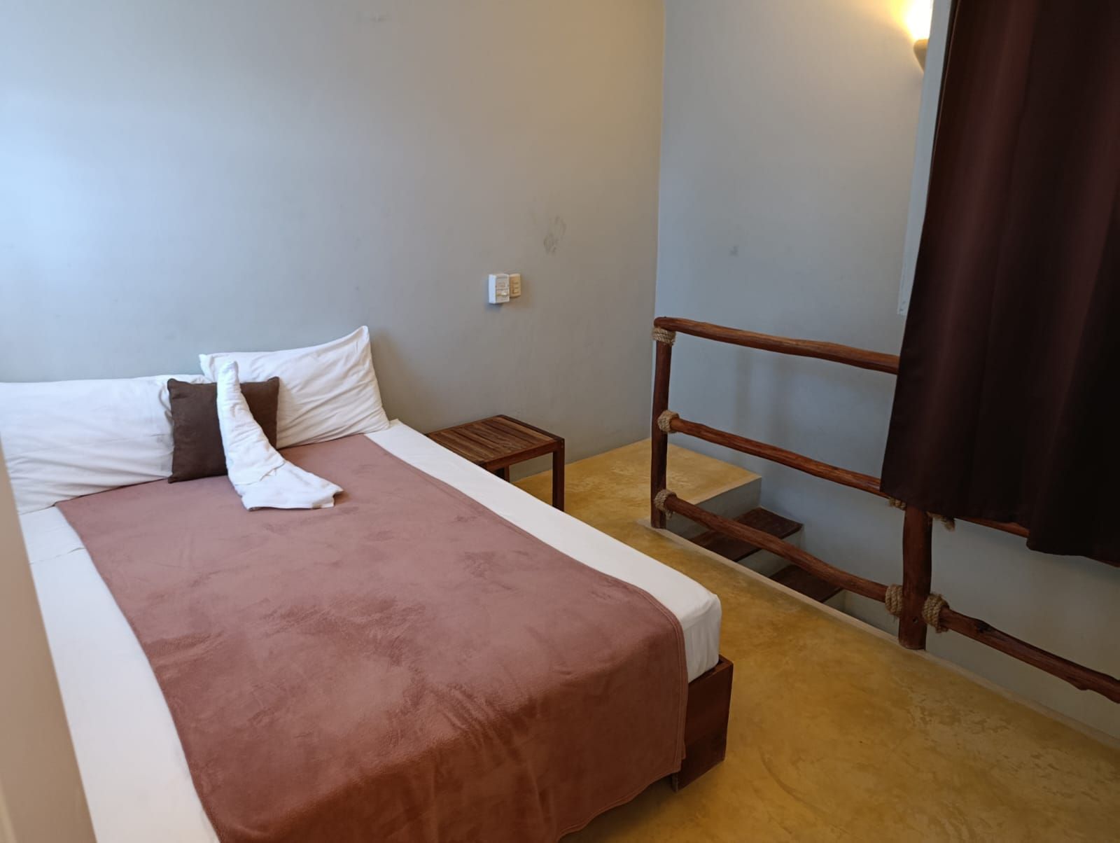 Small hotel room with bed, side table, and stairs to a higher level. Brown and white decor.