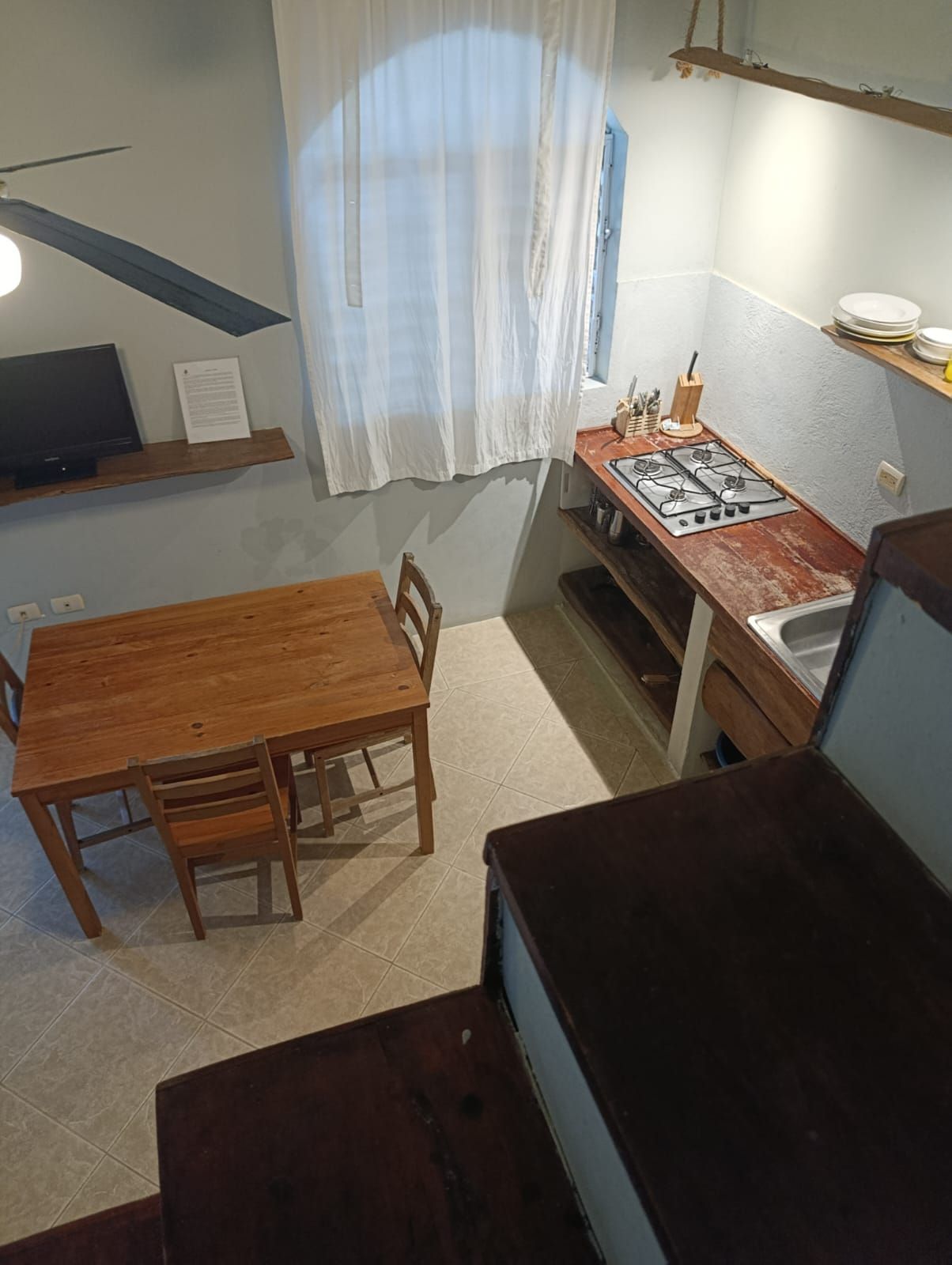 Interior view of small kitchen and dining area with wooden table, gas stove, and white curtain.