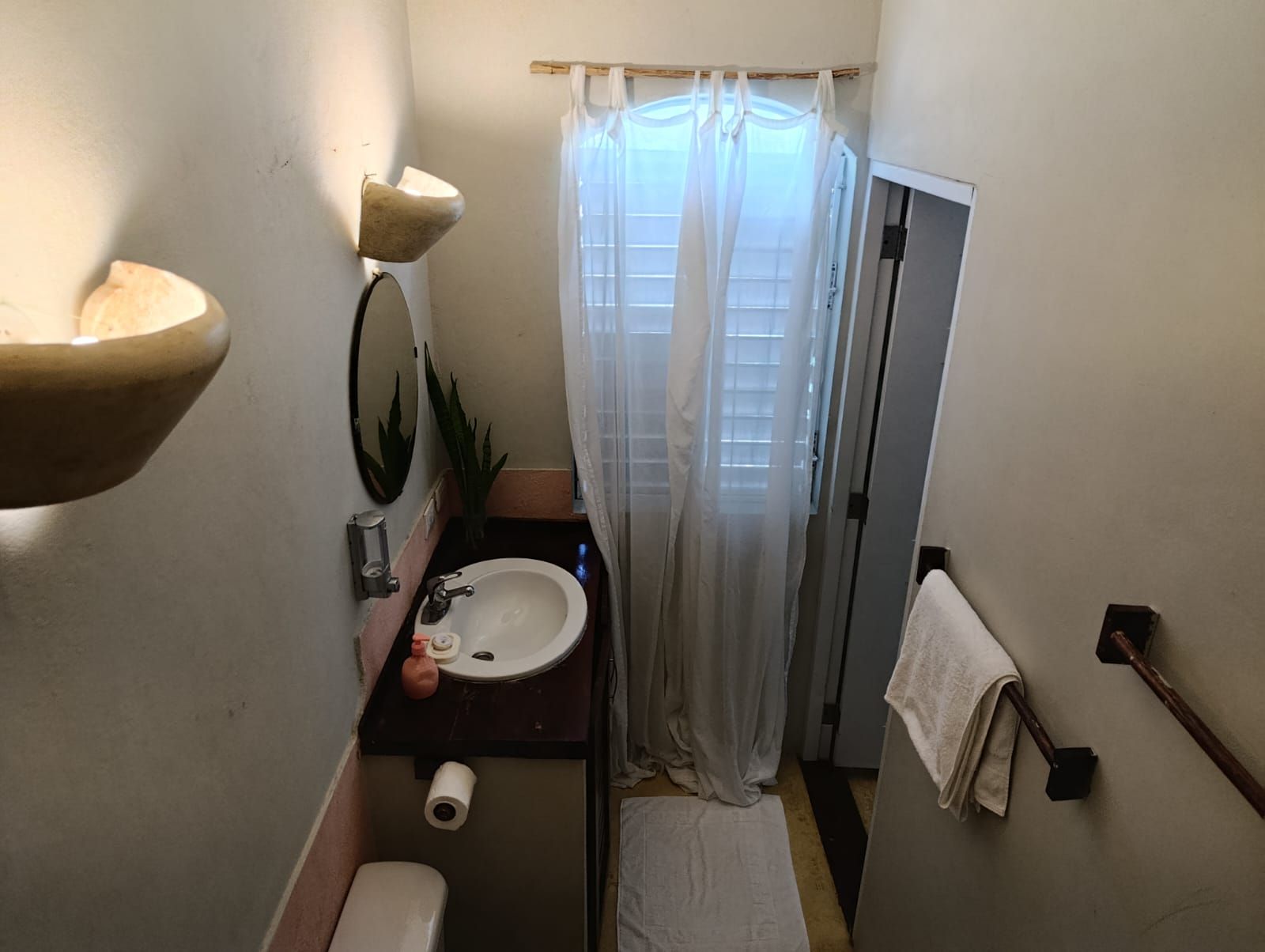 Small bathroom with sink, toilet, and window with sheer curtain. Off-white walls, and earth-toned accents.