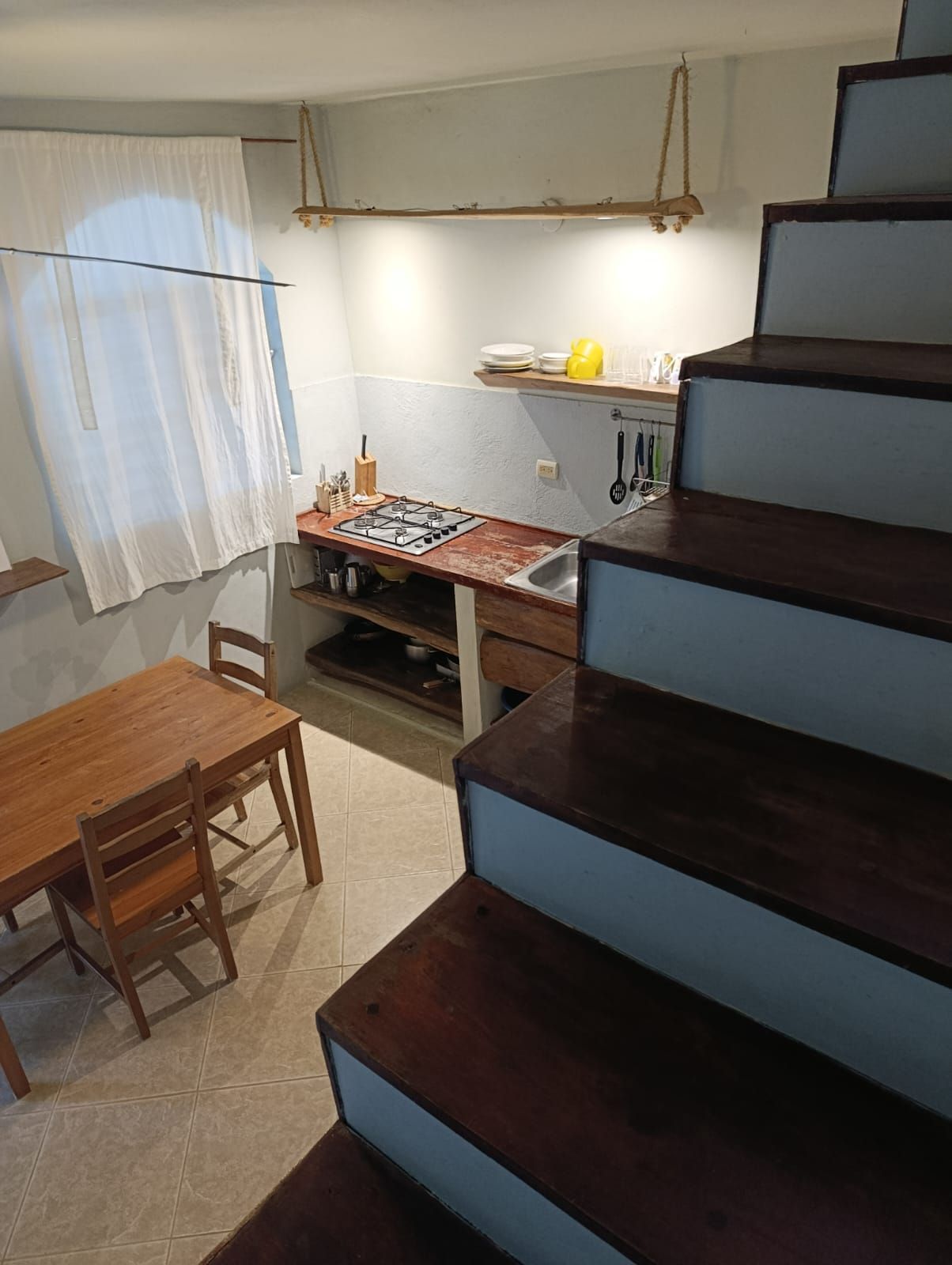 Small kitchen with stairs. Wooden table and chairs on left, stovetop and sink in center, stairs on right.