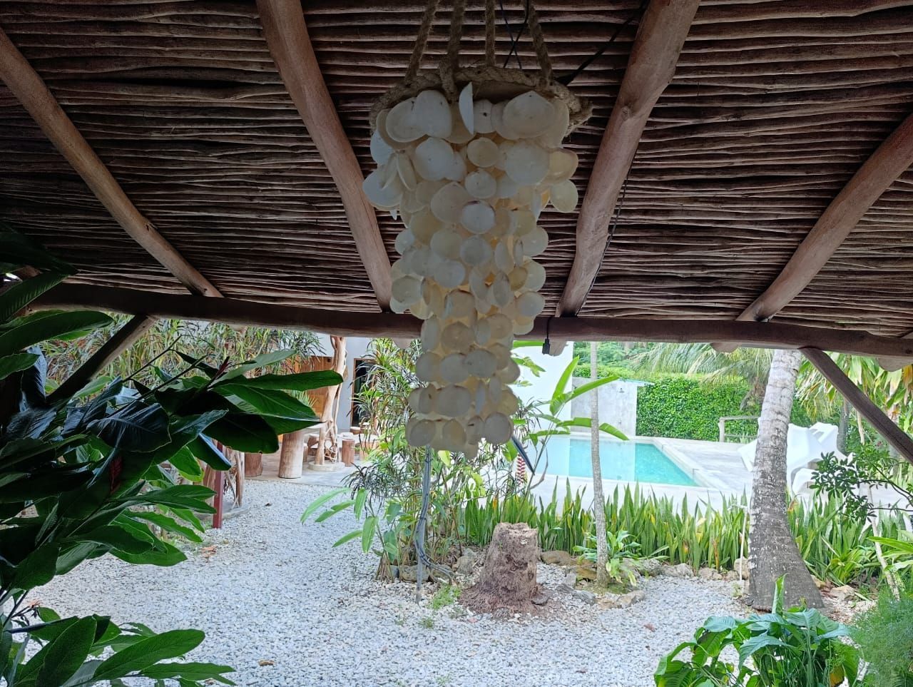 Shell chandelier hanging under a thatched roof, view of a pool and garden.