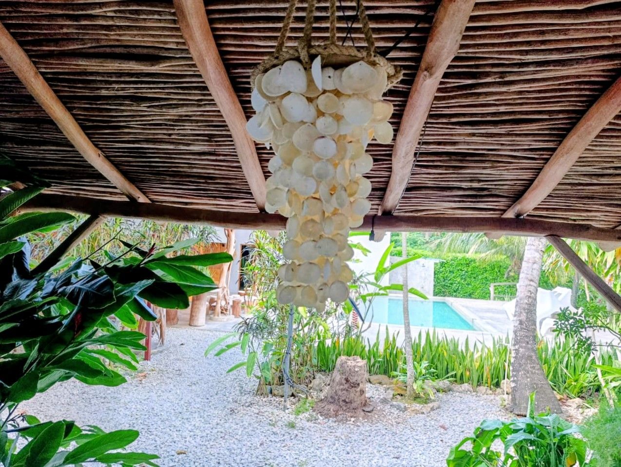 Shell chandelier hangs under a wooden-beamed roof, overlooking a pool and garden.