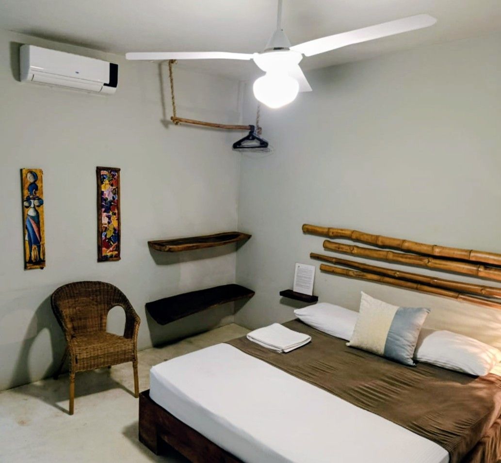 Bedroom with a bed, wicker chair, art, and ceiling fan. Light gray walls, white floor.