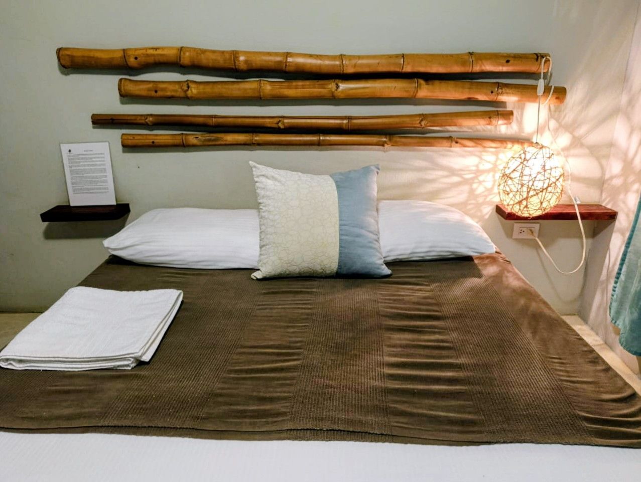 Bedroom with bamboo headboard, brown blanket, white pillows, bedside lamp, and a towel.