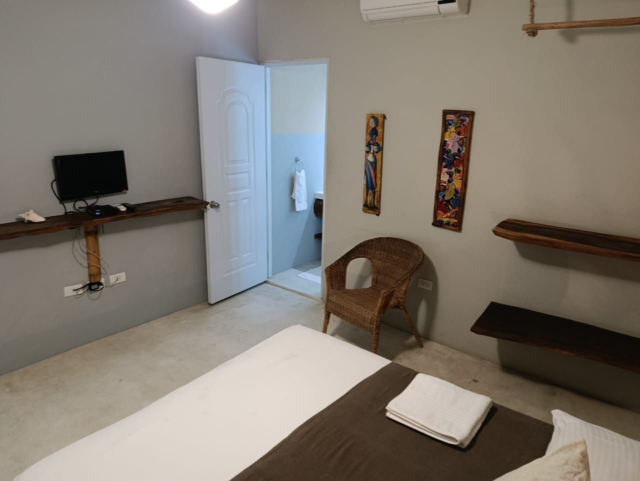 Bedroom with bed, wicker chair, TV, artwork, and open doorway to a bathroom. Gray and white decor.