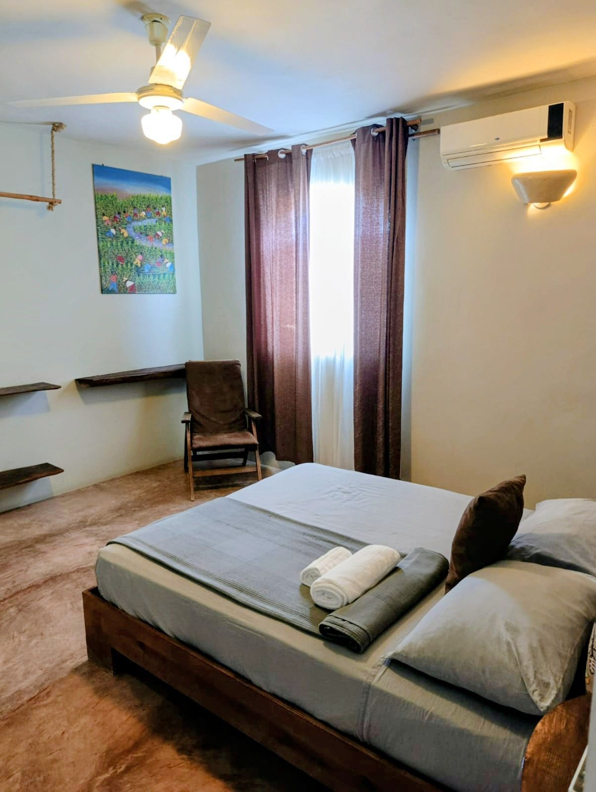 Bedroom with a bed, chair, and art; brown, gray, and white tones; natural light from window.