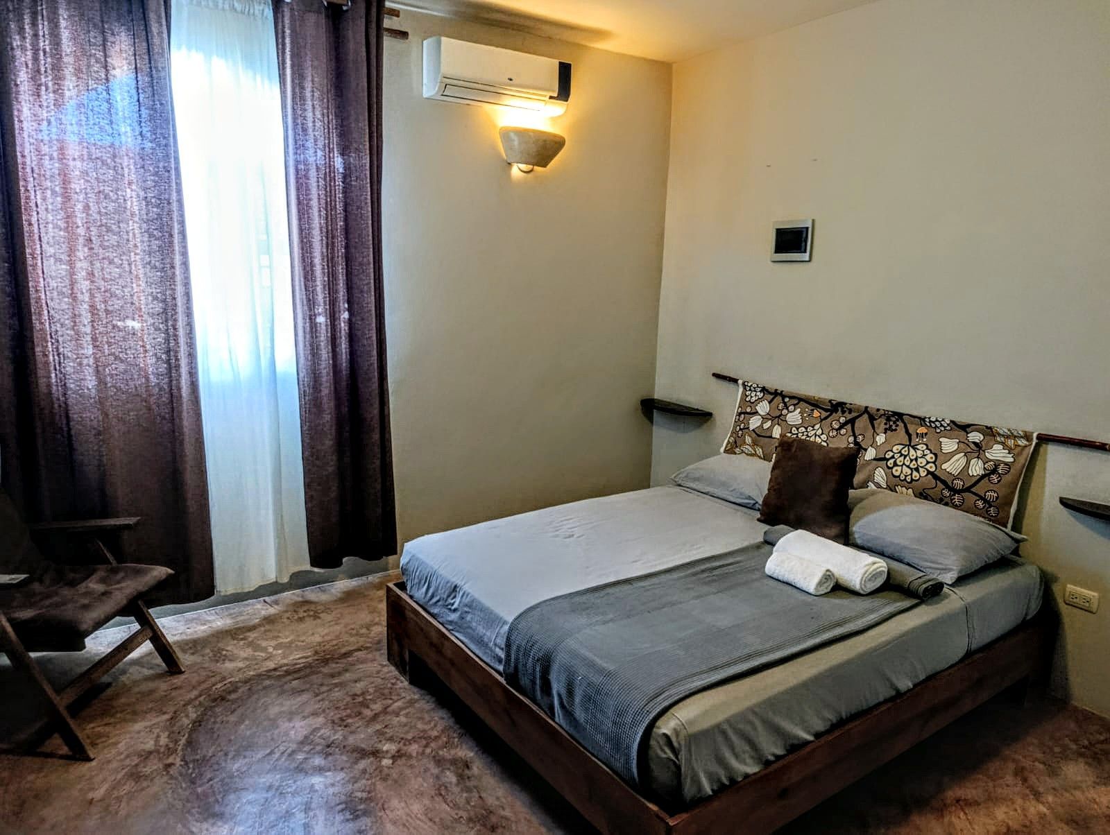 Bedroom with a double bed, dark wood frame, gray bedding, dark brown curtains, and a chair.