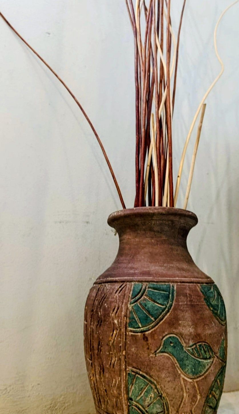 Brown vase with green bird and leaf design, holding tall brown and tan decorative sticks against a white wall.