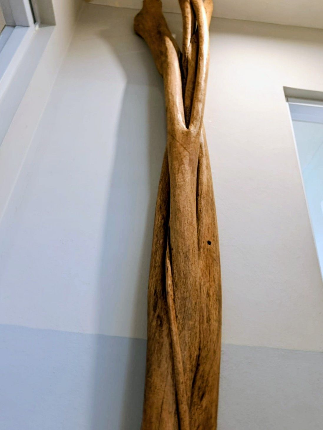 Wooden sculpture against a white and light blue wall. The sculpture is tall, textured, and brown.