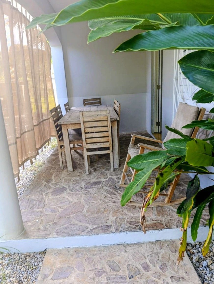 Patio with wooden table and chairs, covered by foliage. Stone floor.