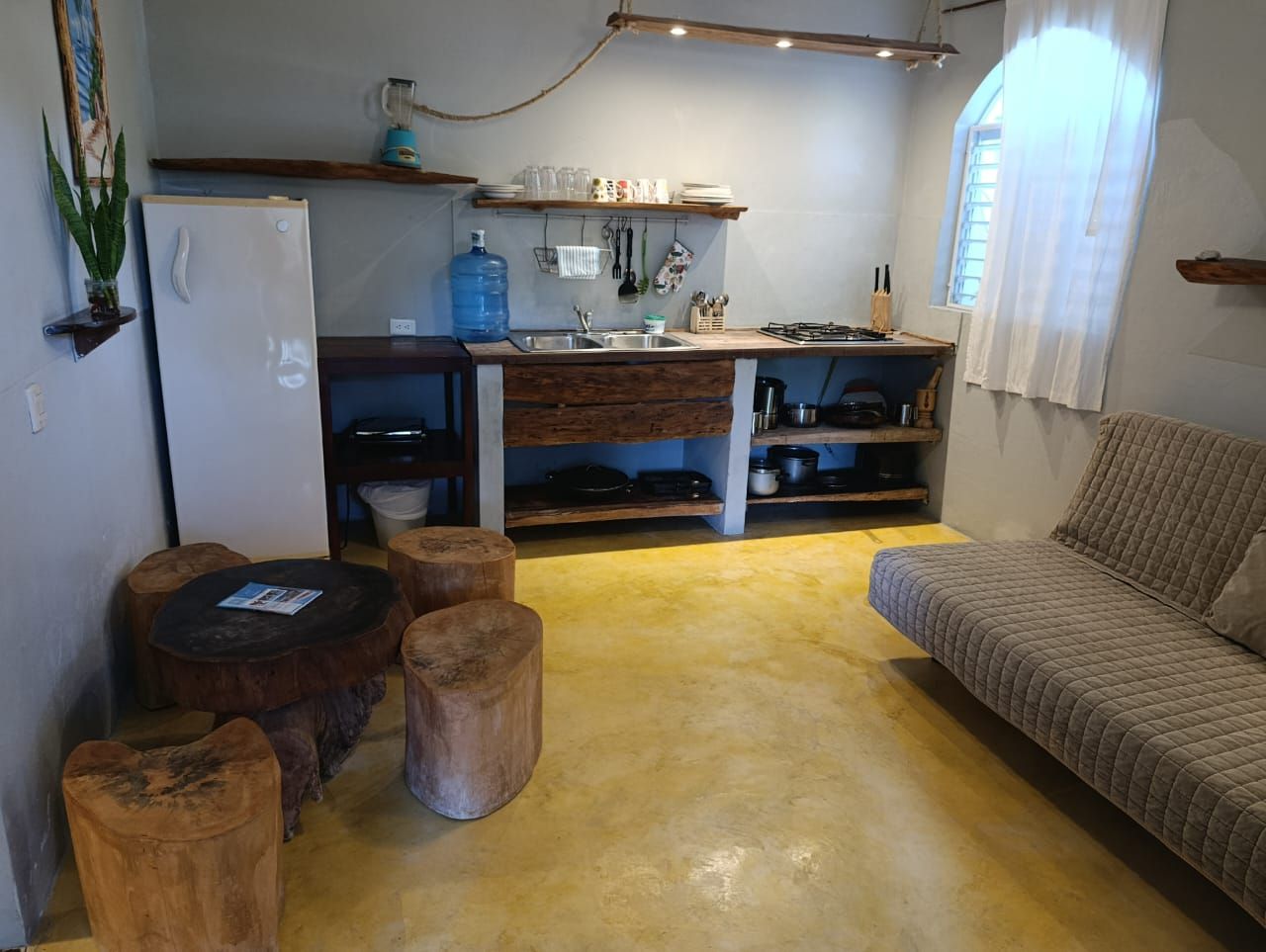A small kitchen and living space with a rustic, wooden aesthetic. Features fridge, sink, and log stools.
