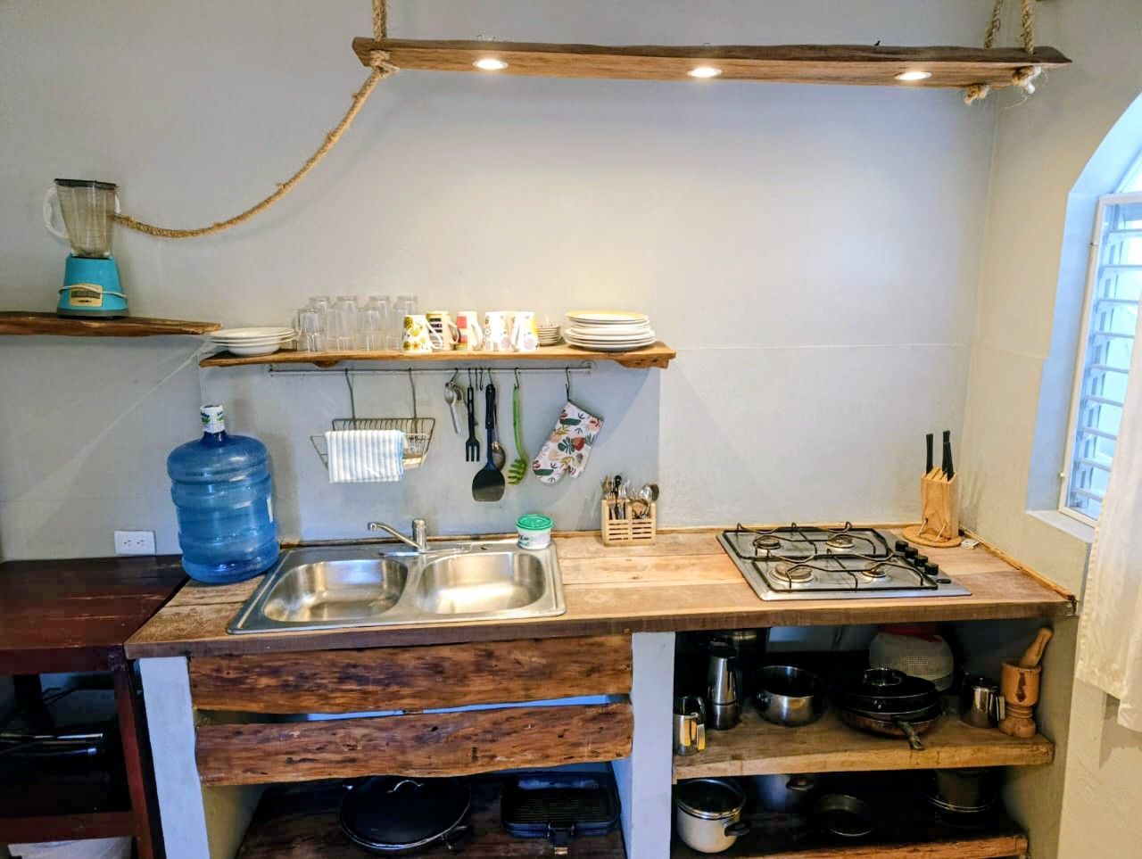 Rustic kitchen with wooden countertops and shelves, two-basin sink, stove, and a large water jug.