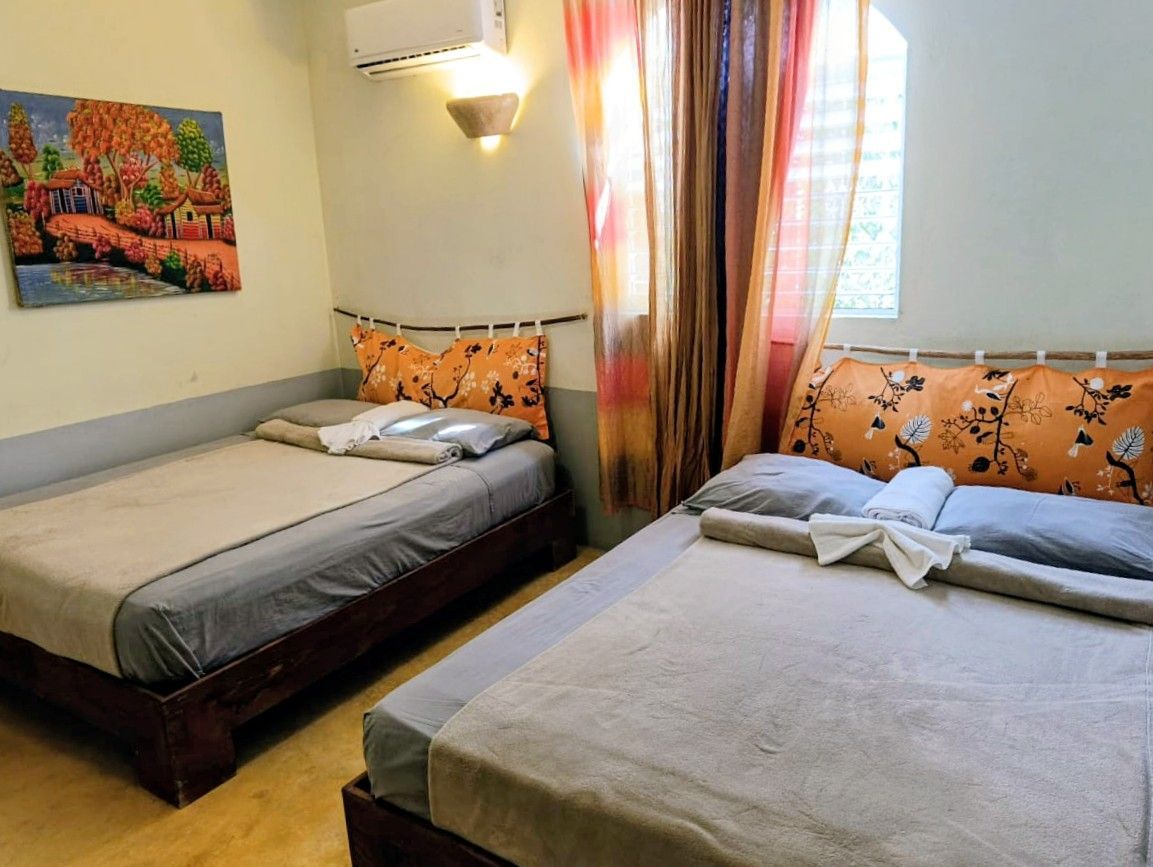 Two beds in a room, with orange and floral pillows. A painting hangs on the wall, curtains frame a window.