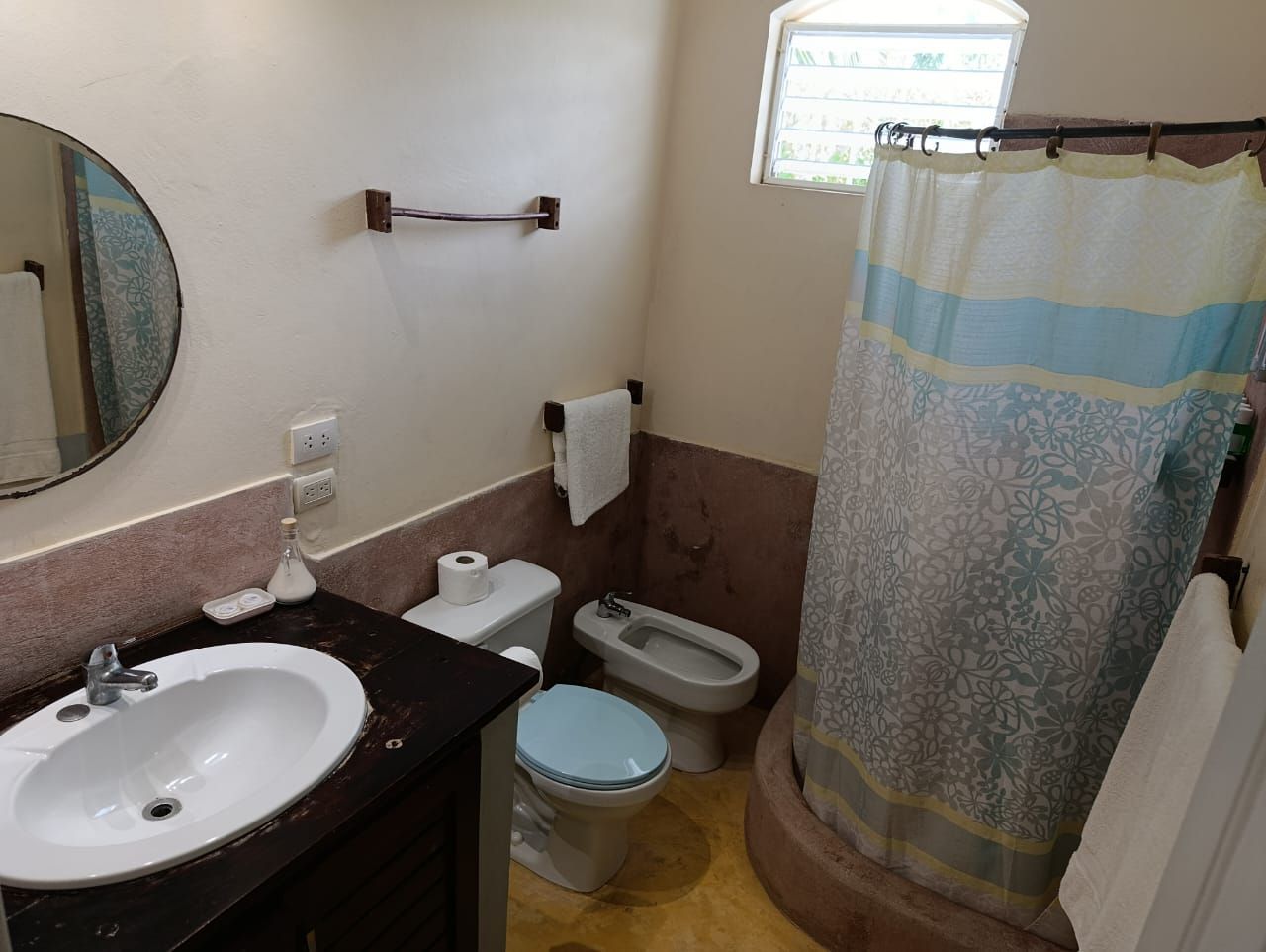 Bathroom with sink, toilet, bidet, shower curtain, and a round mirror. Walls are beige and pinkish-brown.