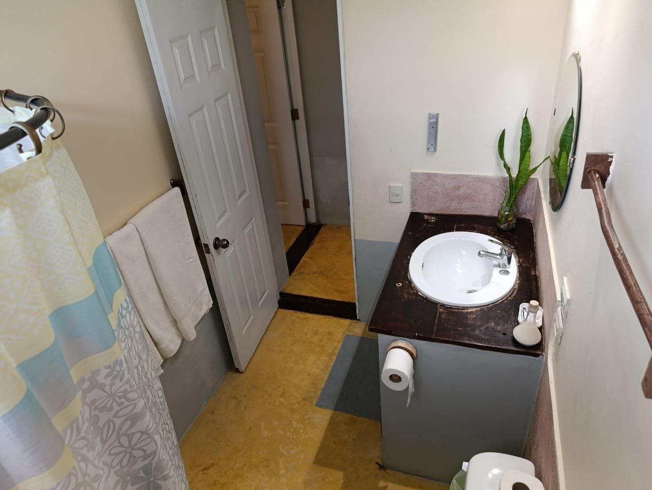 Small bathroom with a sink, toilet, shower, and a doorway. Yellow floor, white door and towels.