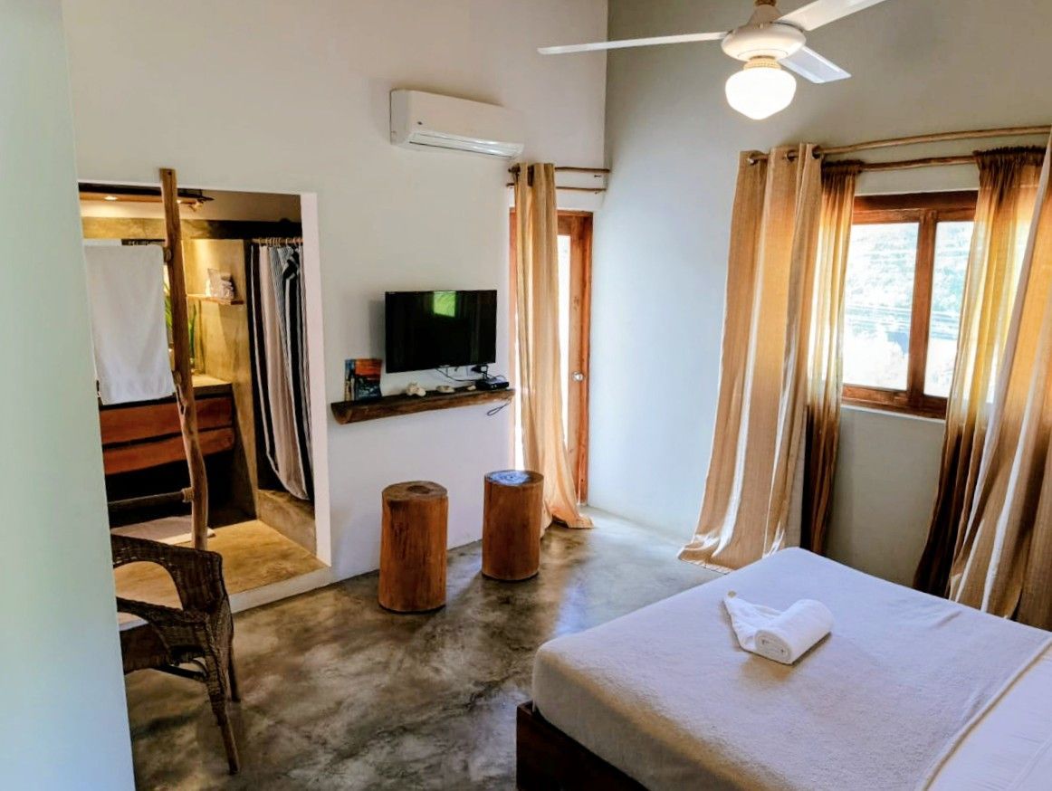 Bedroom with bed, tv, and open bathroom. Light streaming through curtains. Two tree stump seats.