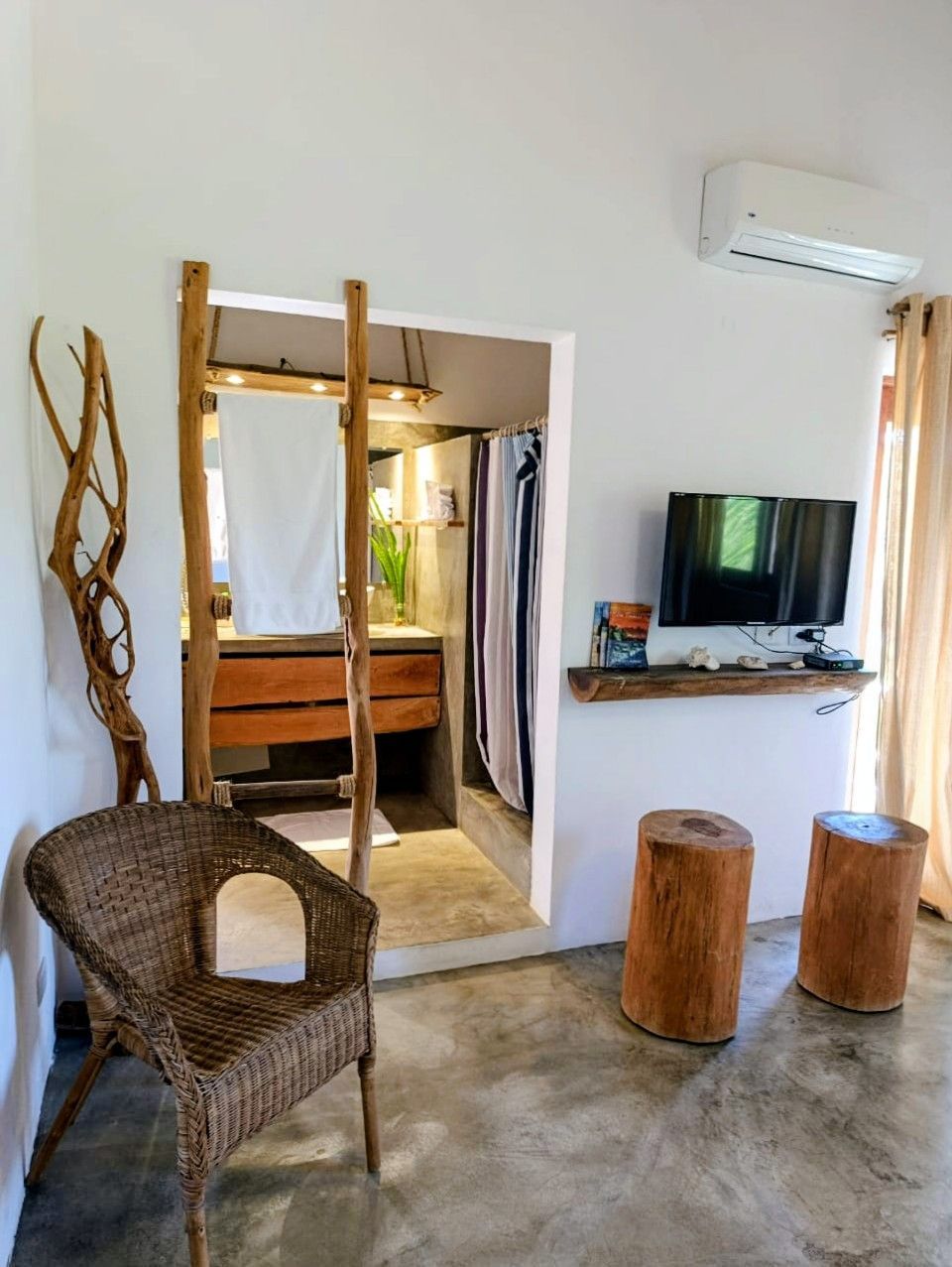 Cozy room with bathroom entry.  Wicker chair, wood stools, mounted TV.  Rustic wood accents, white walls, and concrete floor.