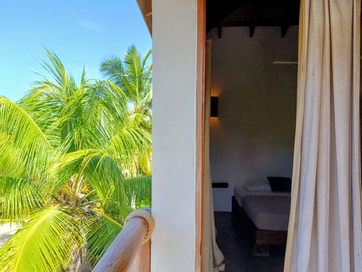 Balcony view: palm trees, white column, interior room with bed, tan curtains, blue sky.