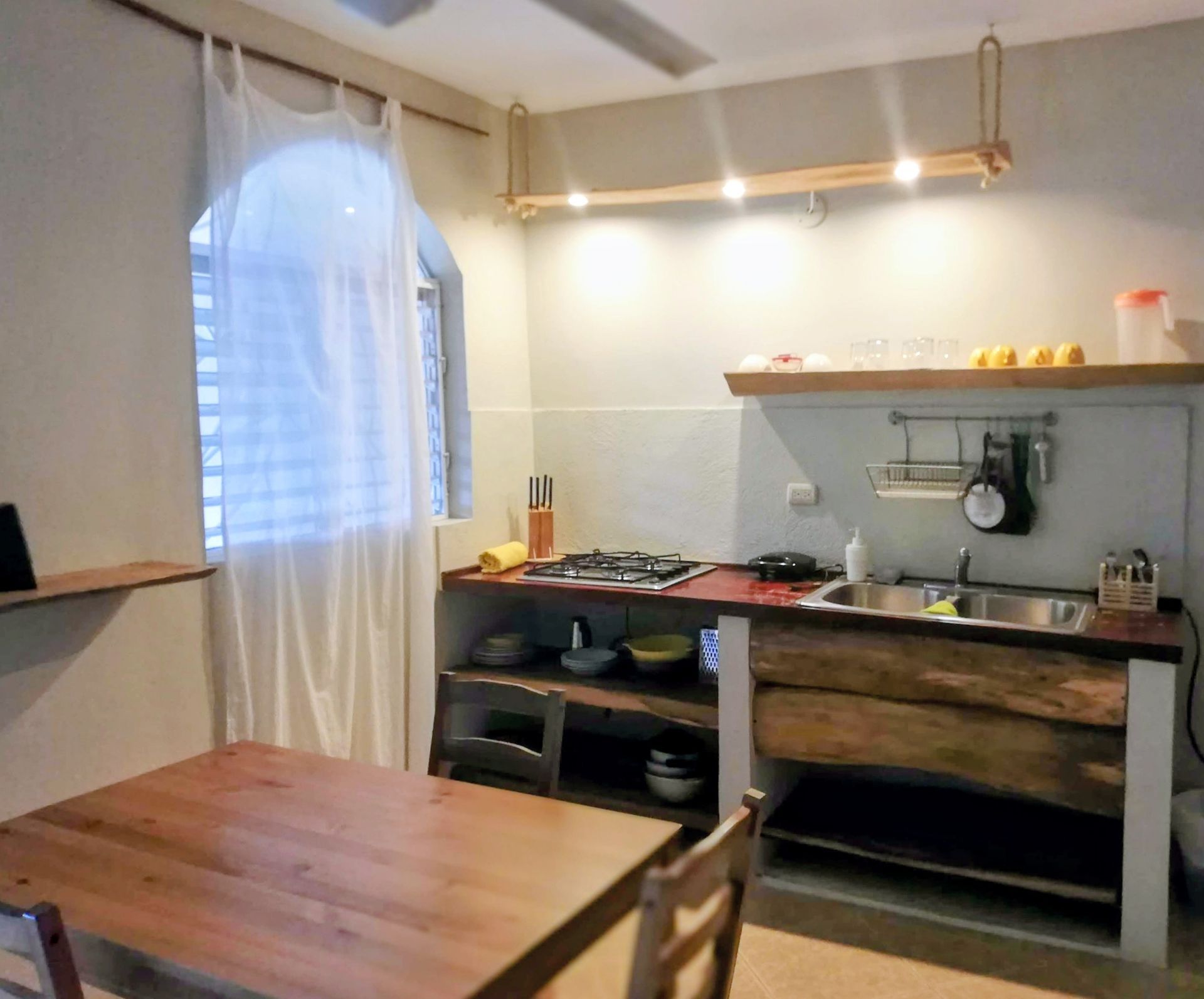 A small kitchen with wood cabinets, red countertop, and a dining table; lit by a window and hanging lights.