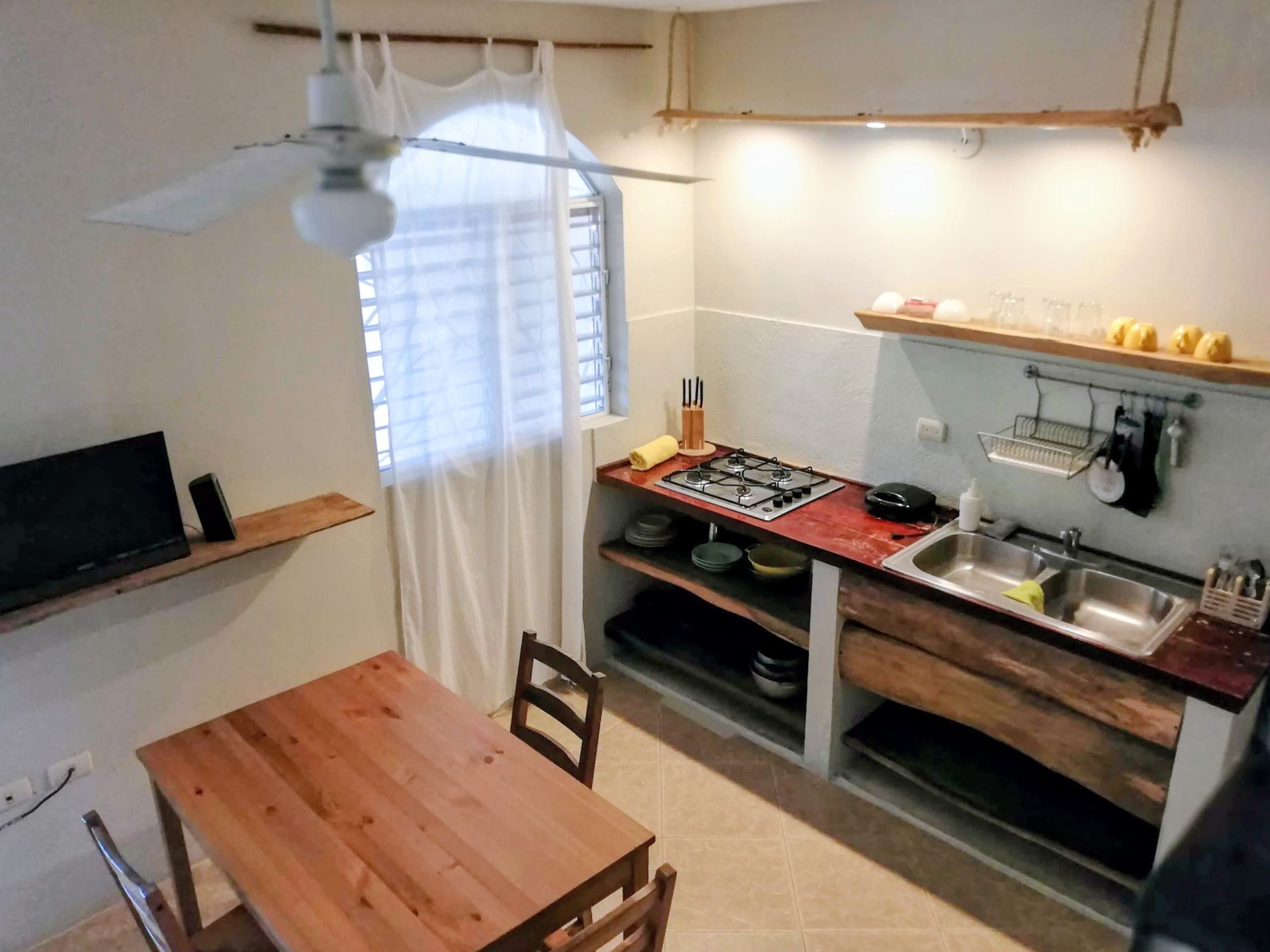 Small kitchen with table and chairs. Stove, sink, shelves, and window with curtain. Wooden accents.