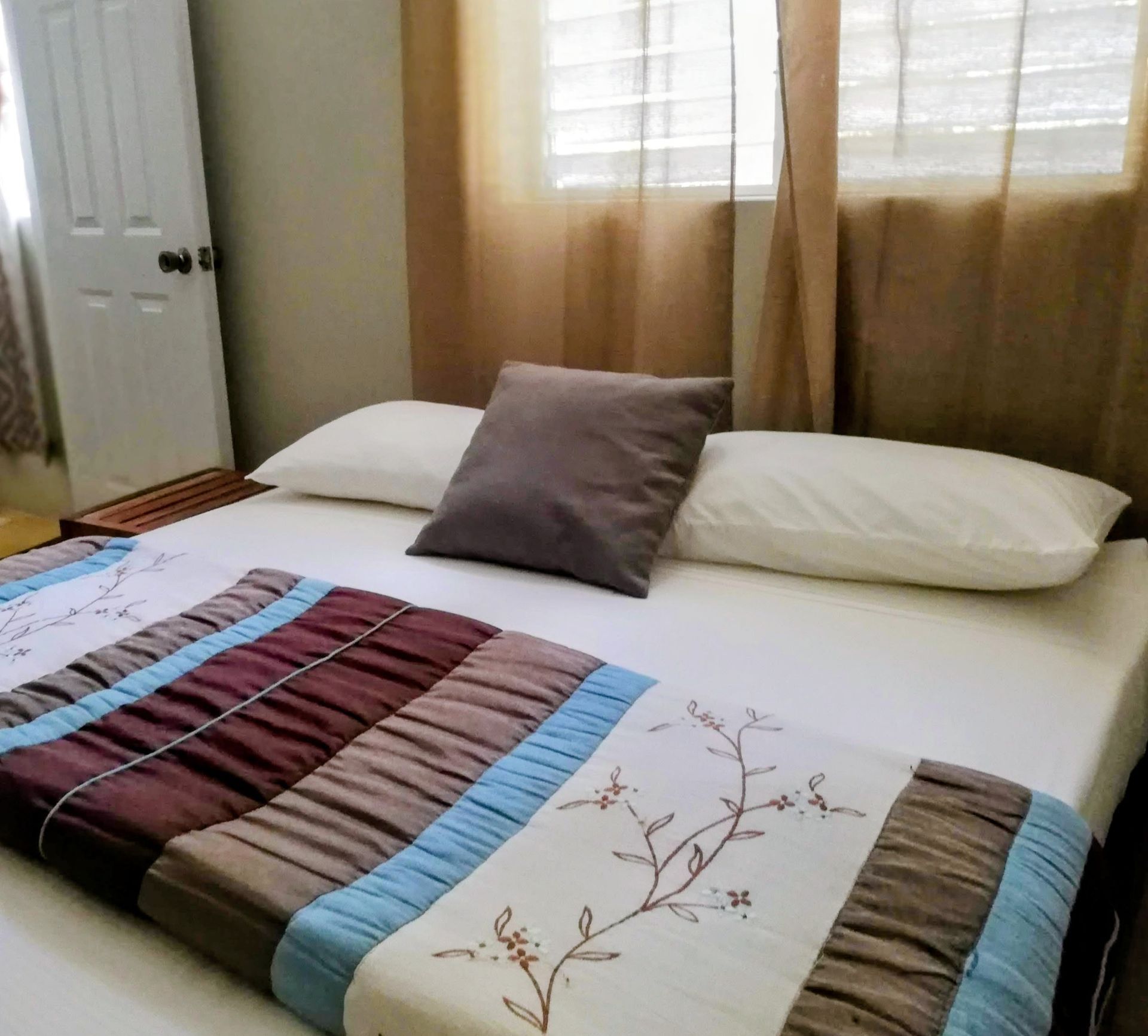 Bedroom with bed, pillows, floral comforter, window with sheer curtains.