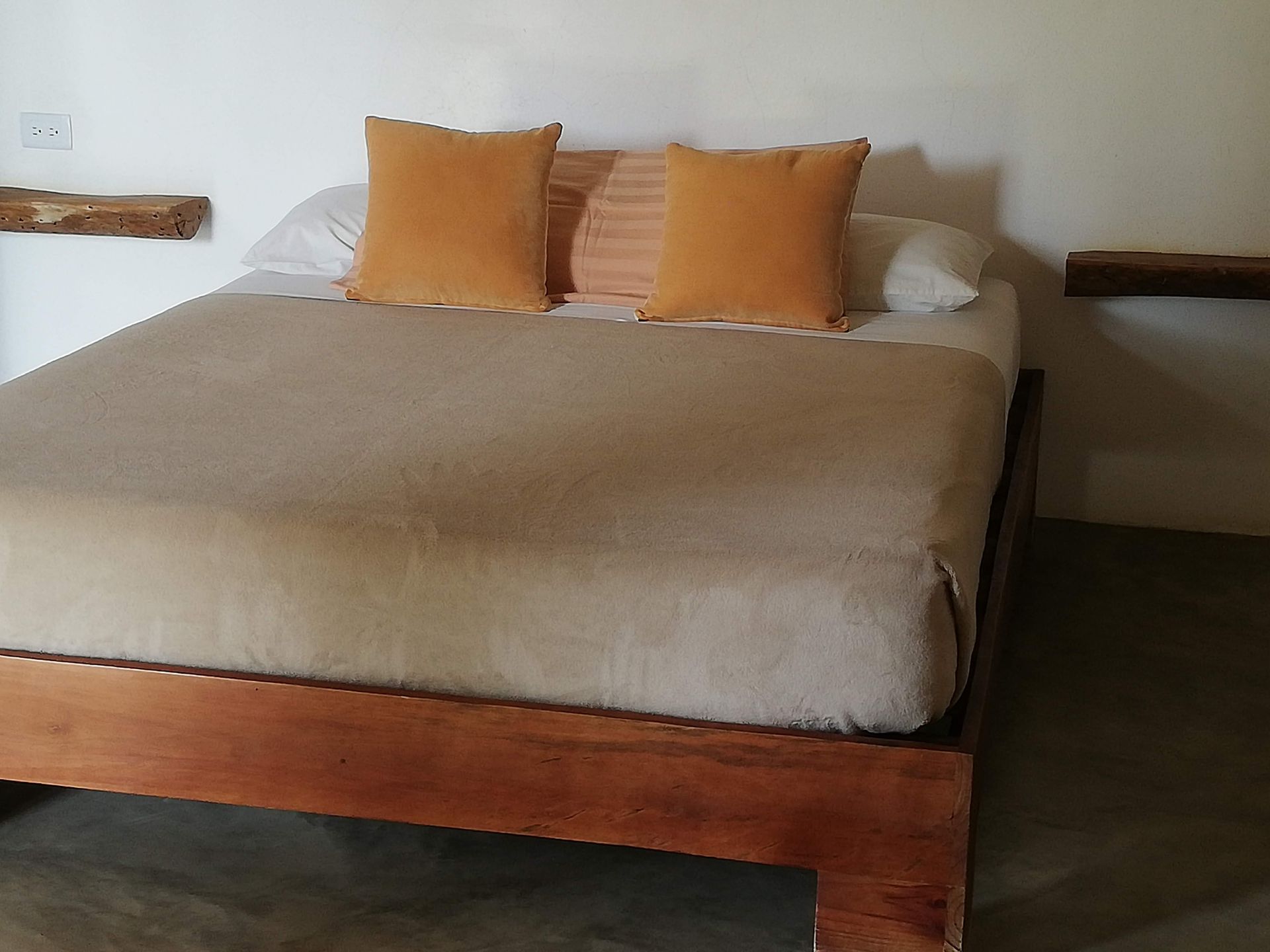 Bedroom with wooden bed frame, tan bedding, orange pillows, and small wooden shelves.