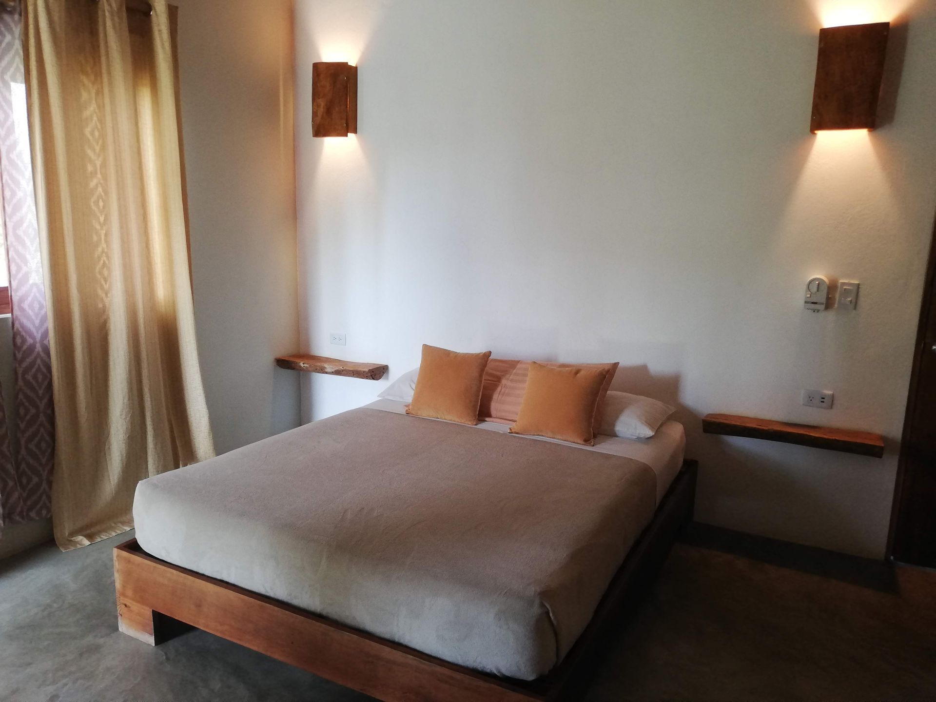 Bedroom with wooden bed, nightstands, and wall sconces. Beige bedding and orange pillows.