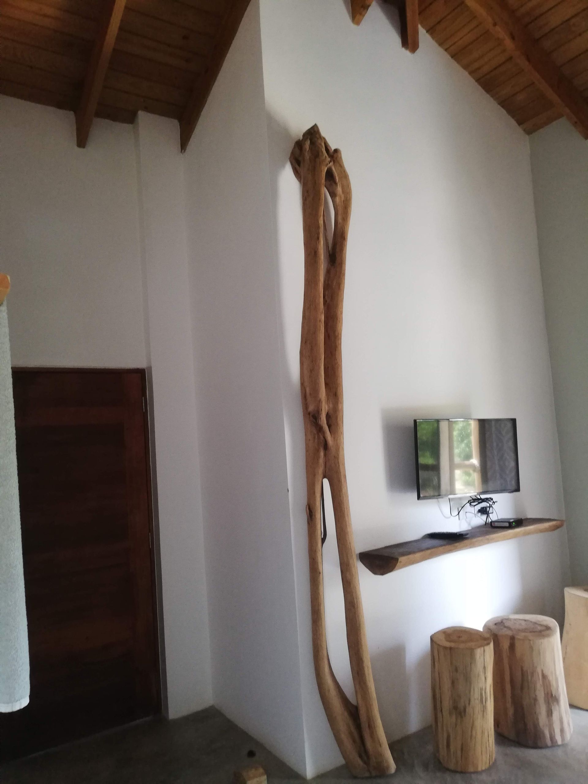 Tall, weathered wood sculpture against white wall in a room; TV and wooden stools.