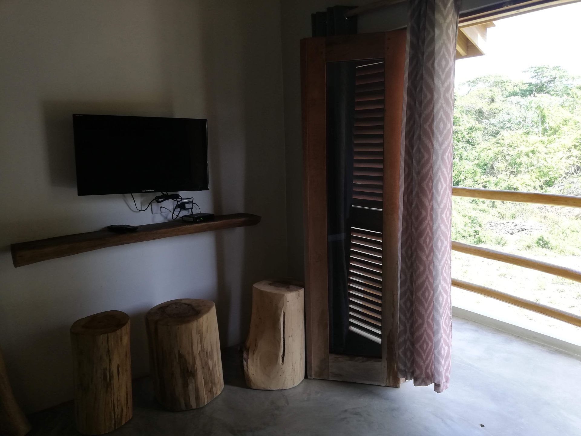 Room corner with a TV, wooden stools, and a balcony overlooking greenery.