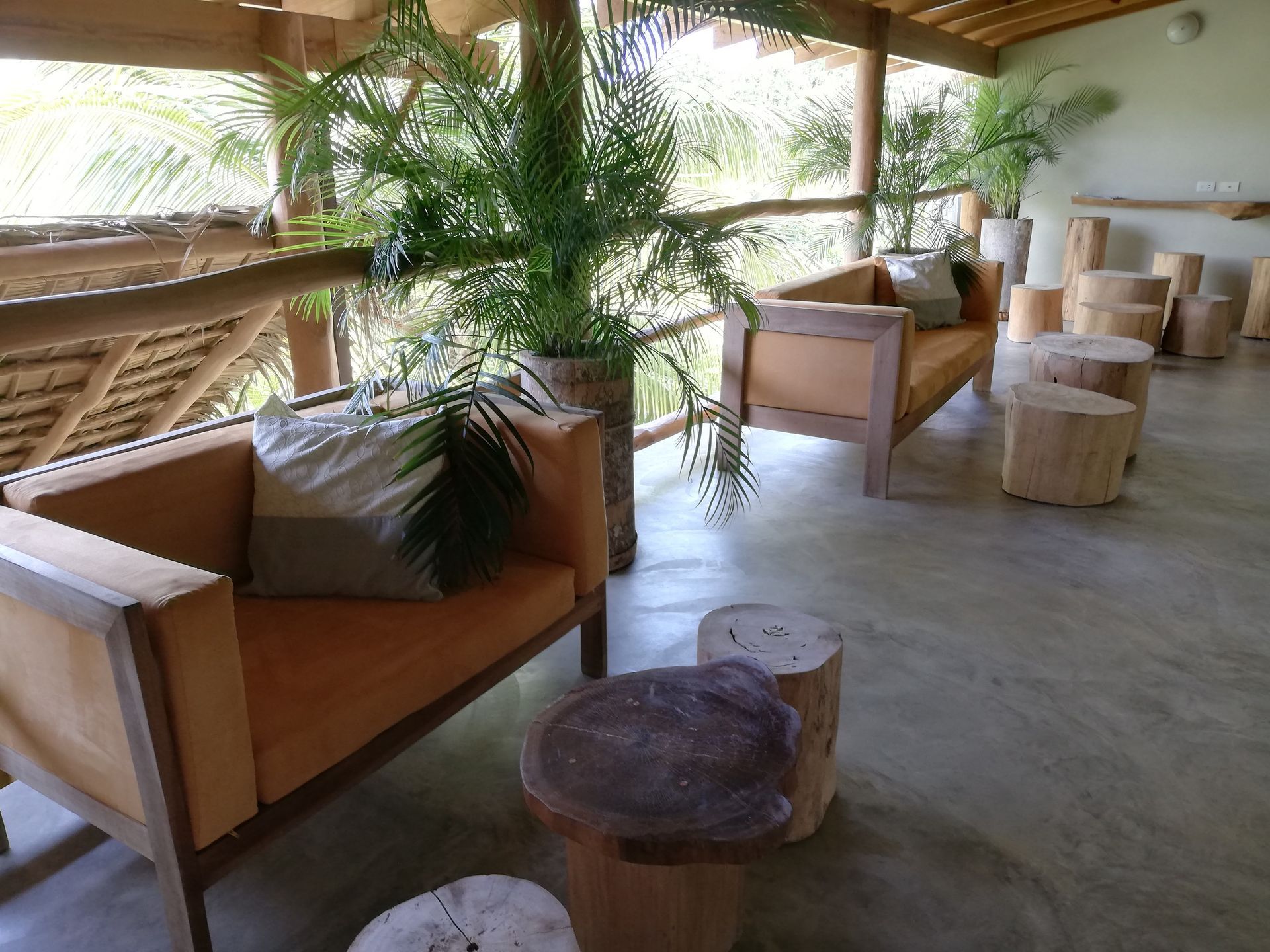 Lounge area with orange couches, wooden side tables, and potted plants on a patio with a wooden railing and ceiling.