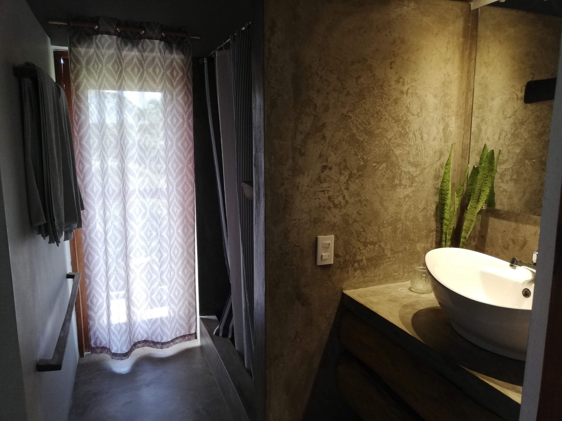Bathroom with a concrete wall, white sink, and sheer patterned curtain; natural light coming in.