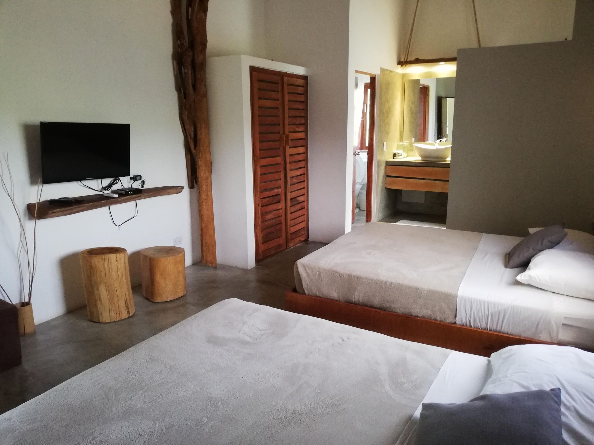 Bedroom with two beds, TV, wooden closet, and bathroom. Concrete floor, neutral colors, rustic style.