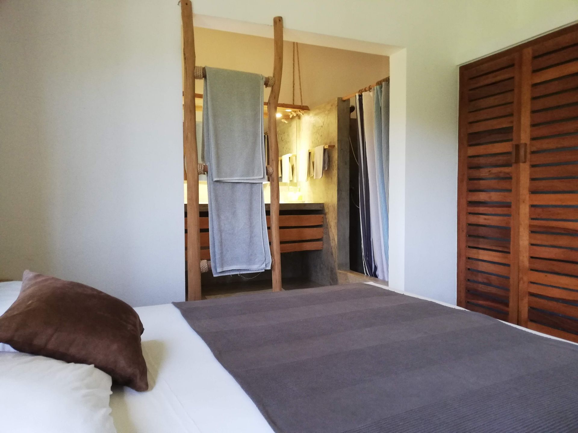 Bedroom with bed, bathroom in the background, wooden wardrobe and towel rack, white walls, and neutral colors.