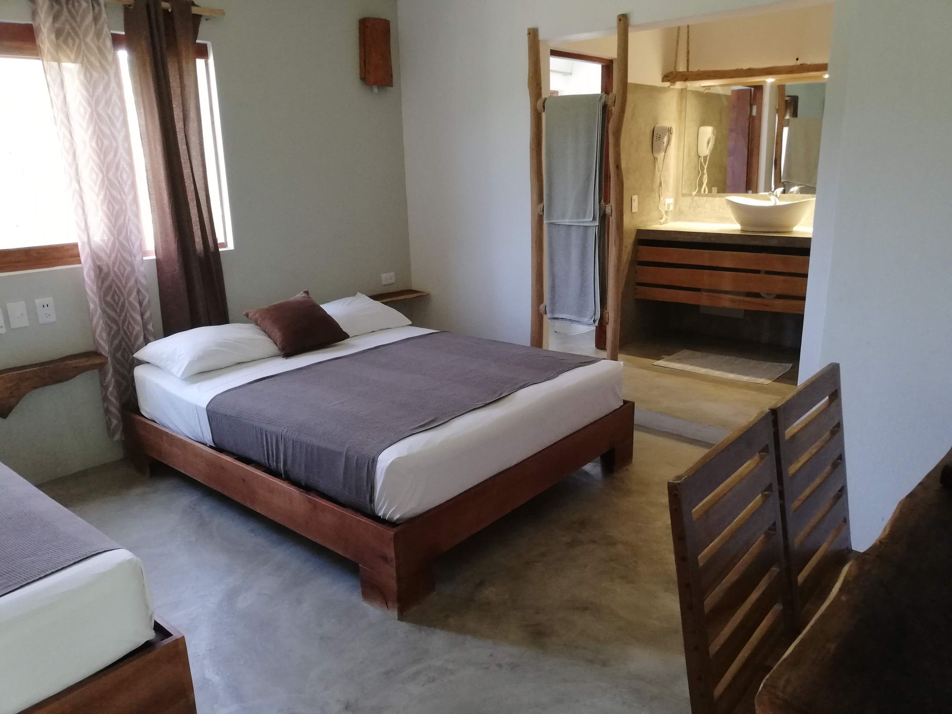 Bedroom with two beds, gray comforters, a window with brown curtains, and a partially visible bathroom.