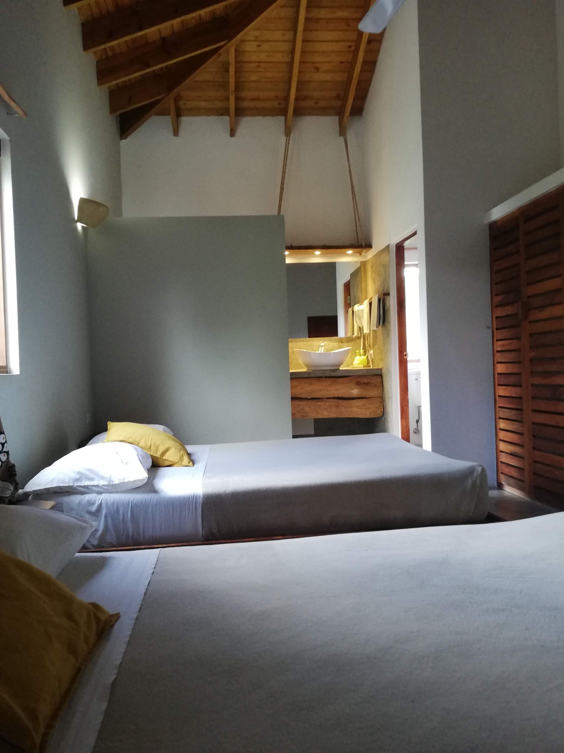 Two beds in a room, a half wall separates the beds from a bathroom. Light wooden ceiling.