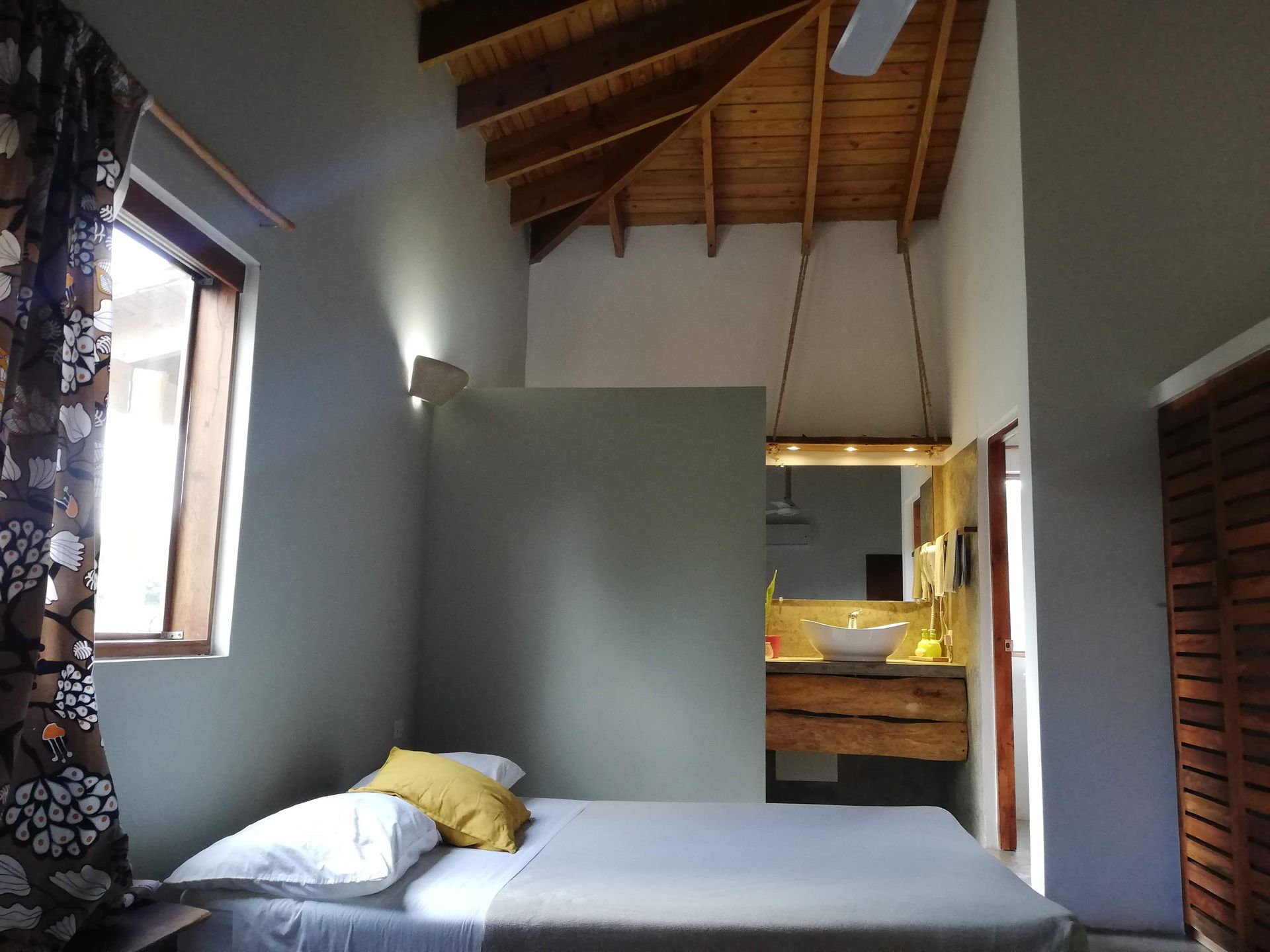 Bedroom with bed, bathroom in background, high wood ceiling, window with floral curtain.