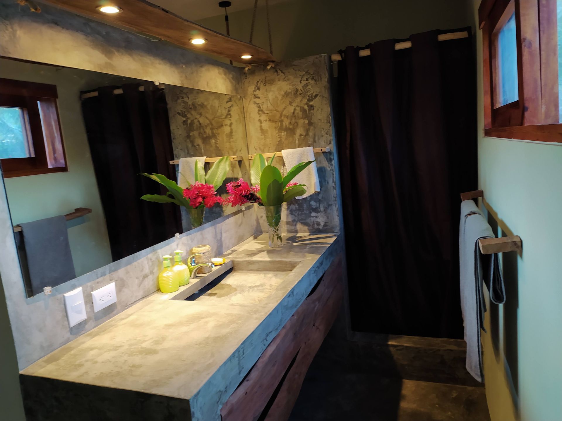 Bathroom with a concrete countertop, large mirror, and dark curtains. Features flowers and wooden accents.