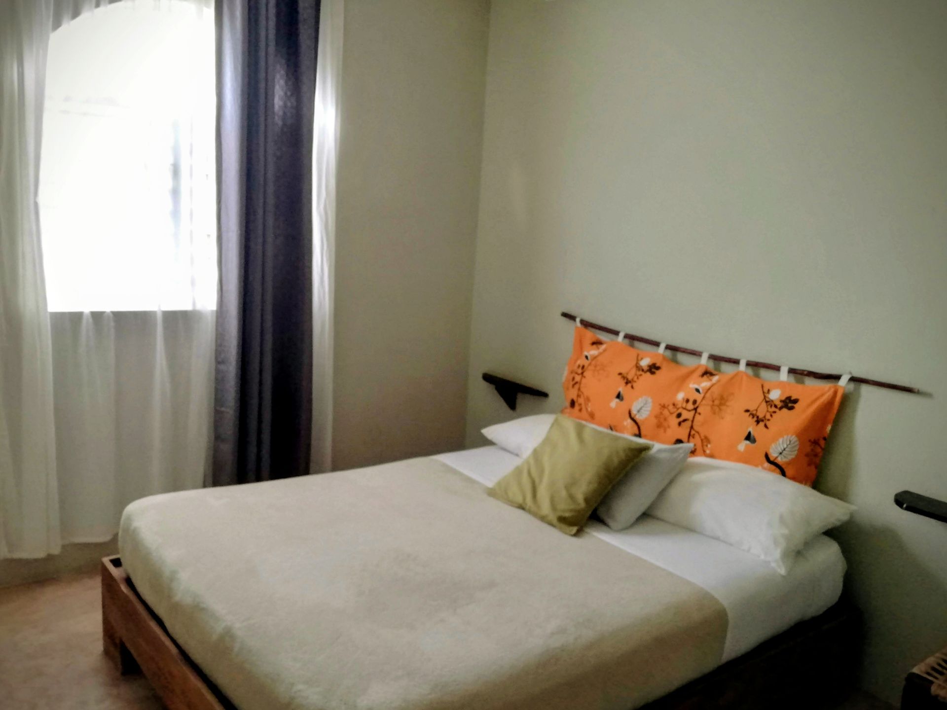 Bedroom with double bed, pillows, window with curtains. Bed has an orange headboard.