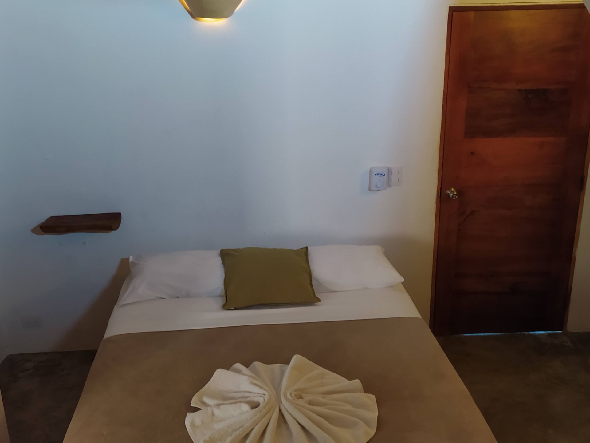 Small, simple bedroom with a bed, white walls, wooden door, and a shelf.