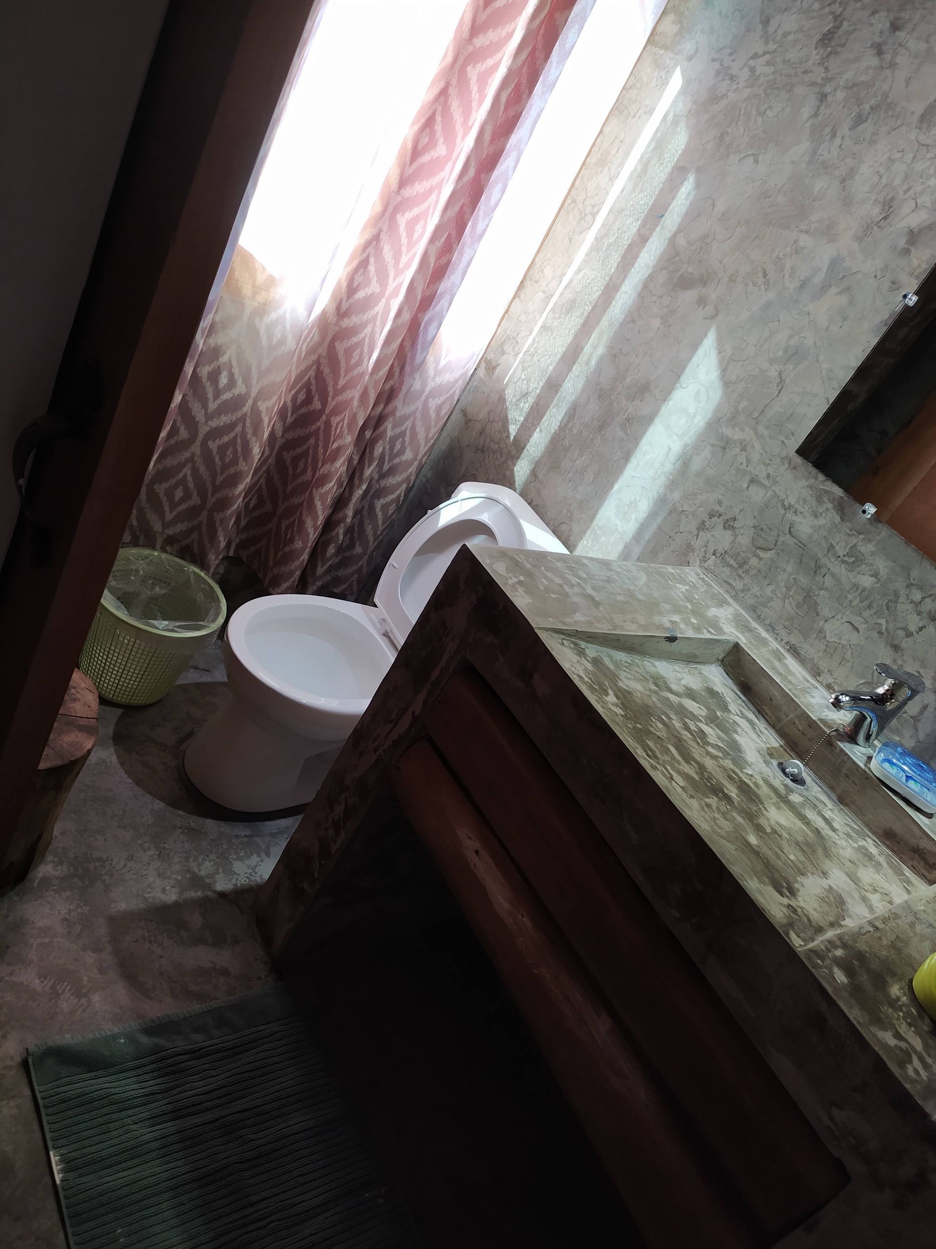 Bathroom with toilet, sink, window, and partially closed curtain. Sunlight streams in.