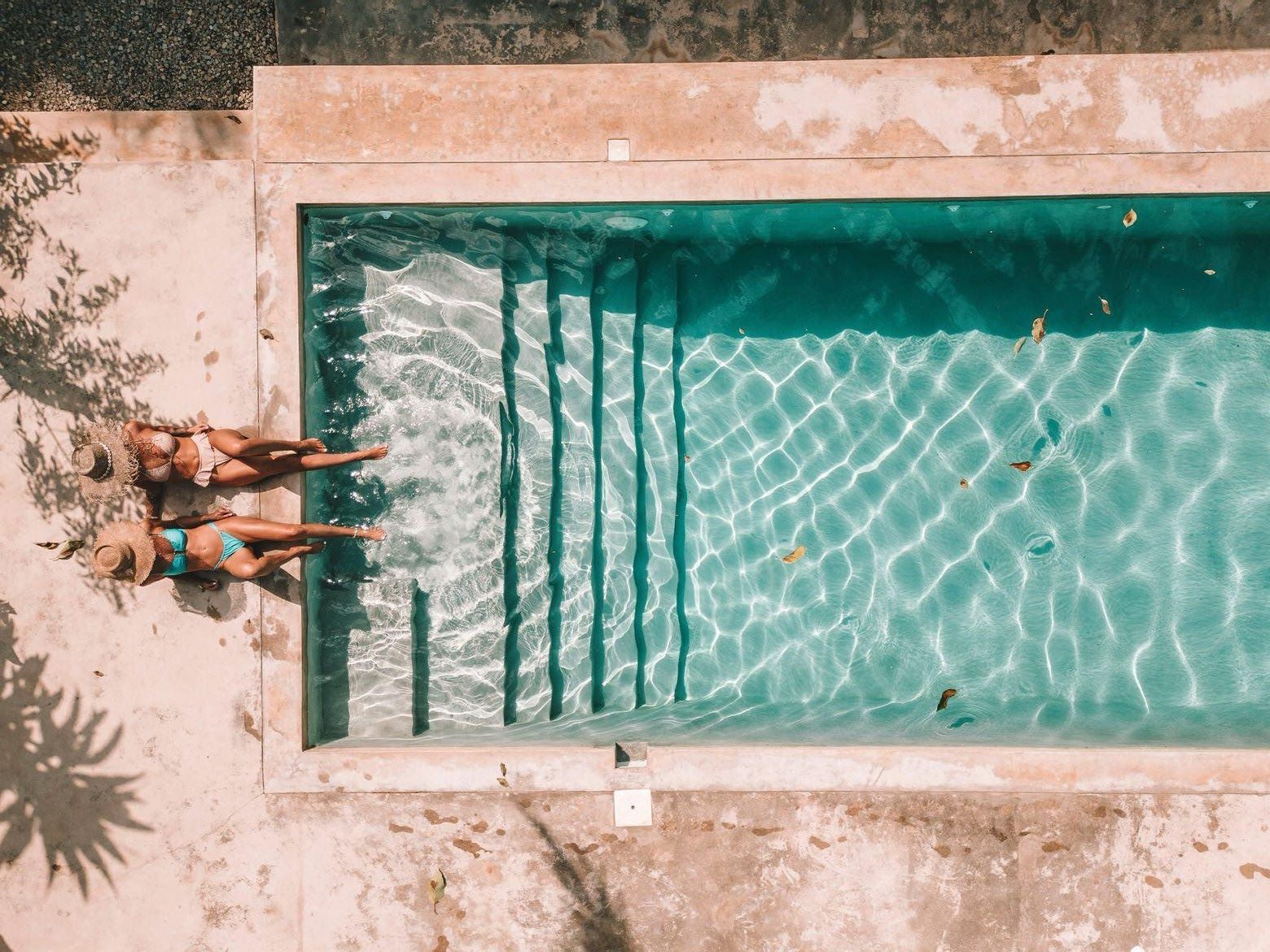 Two women sunbathing by a turquoise pool with steps, viewed from above.