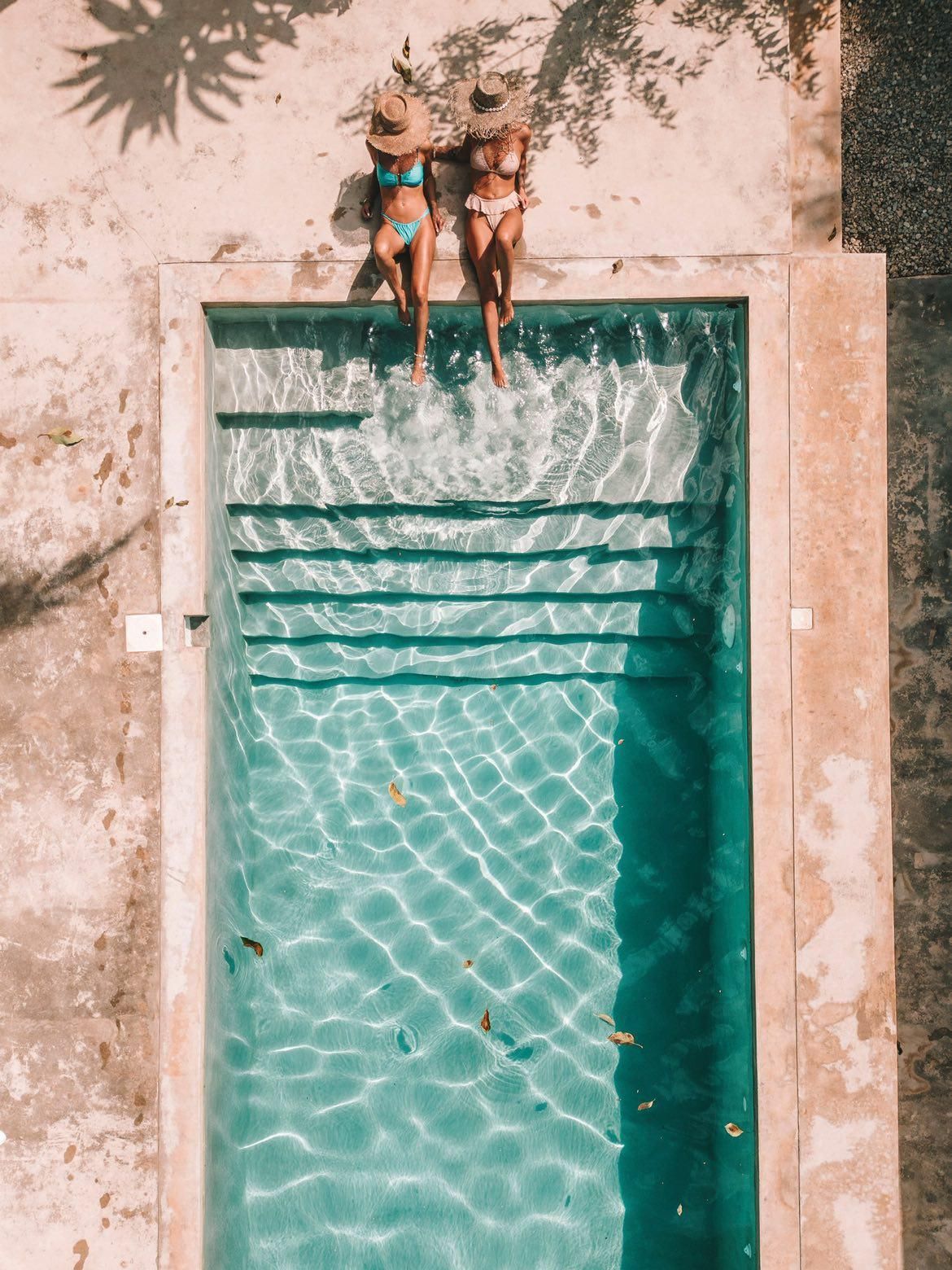 Two women in swimsuits sunbathe by a turquoise pool. Sunlight casts shadows on the aged concrete.