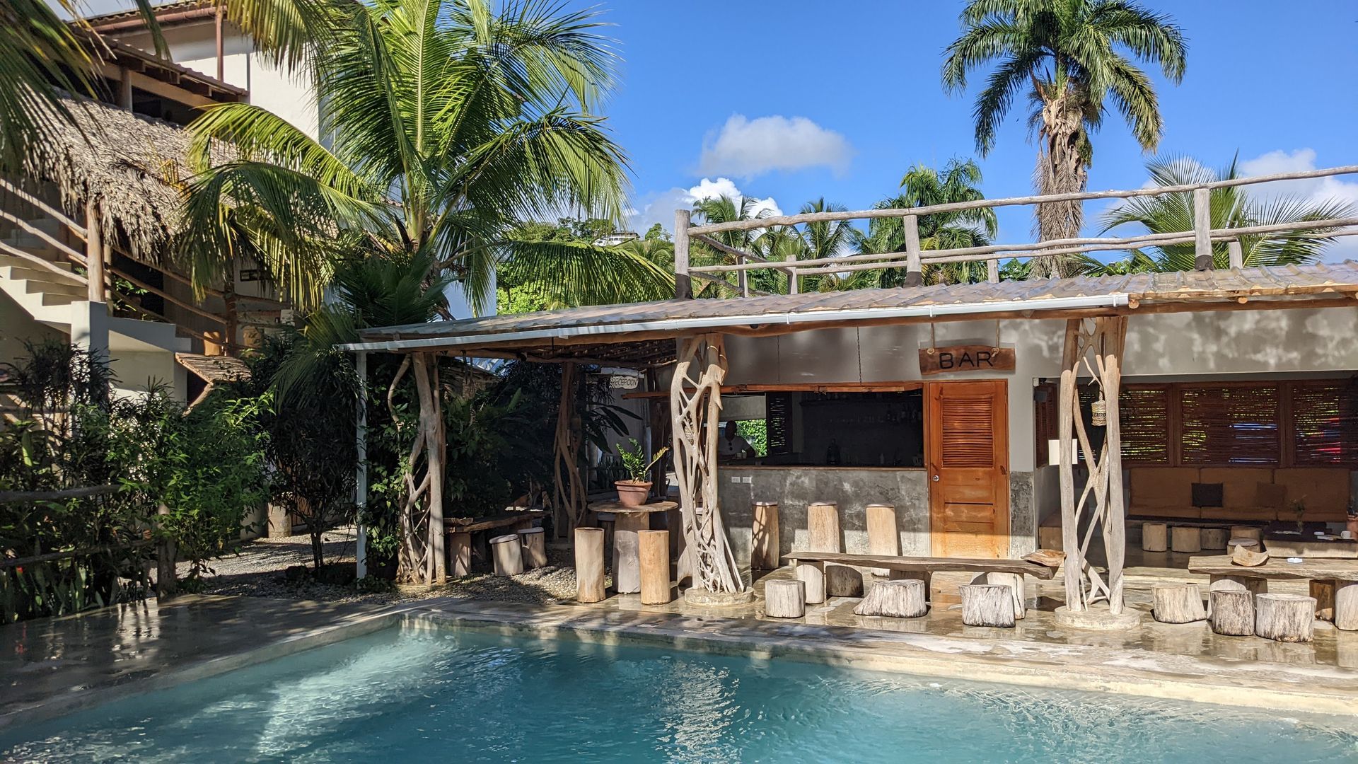 Poolside bar with thatched roof and wooden supports, next to a blue pool, under a bright blue sky.
