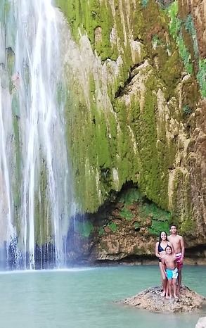 Family stands on a rock at a waterfall. Bright blue water, green mossy cliffs.