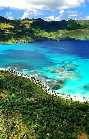 Turquoise bay with a sandy beach lined with palm trees, and green hills under a blue sky.