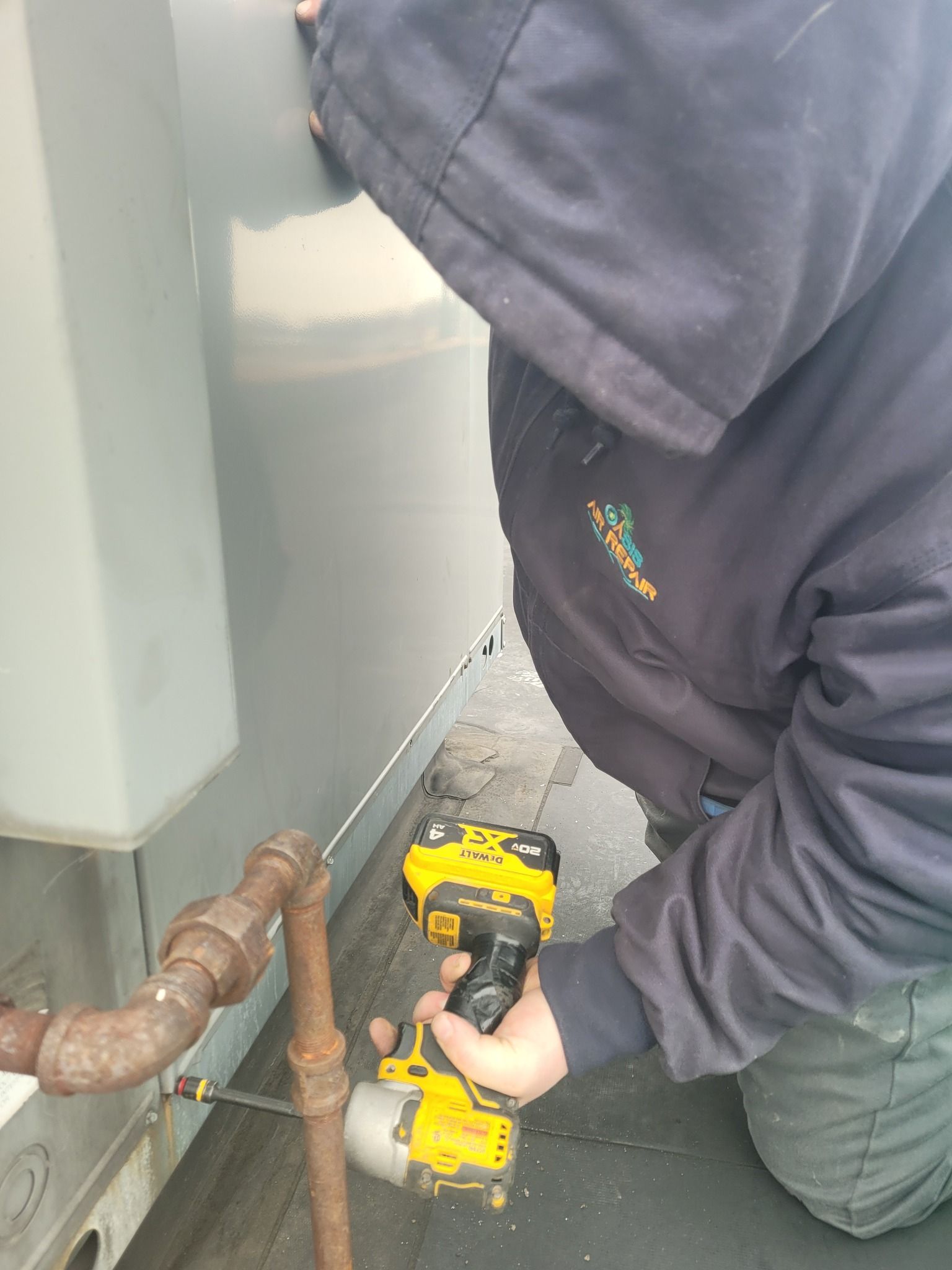 A person in a dark hooded jacket kneeling on a roof uses a yellow power drill on a metal gas pipe and panel.