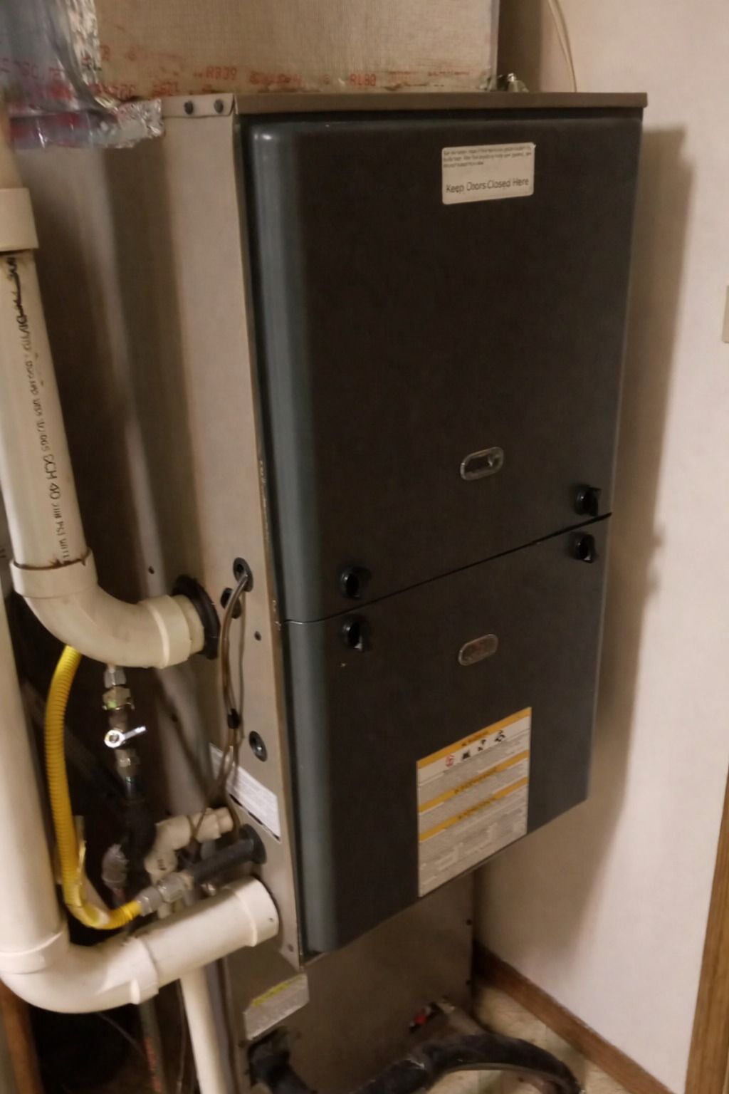 A vertical, dark grey residential furnace unit with white PVC piping attached to the side, mounted in a room.