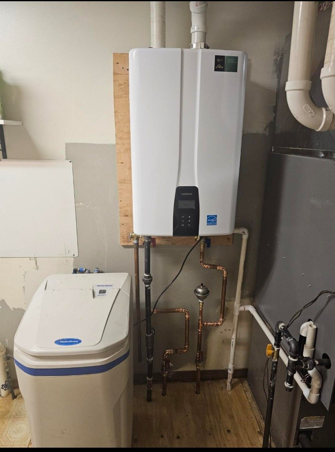 A white tankless water heater mounted on a wooden board above copper plumbing pipes next to a water softener unit.