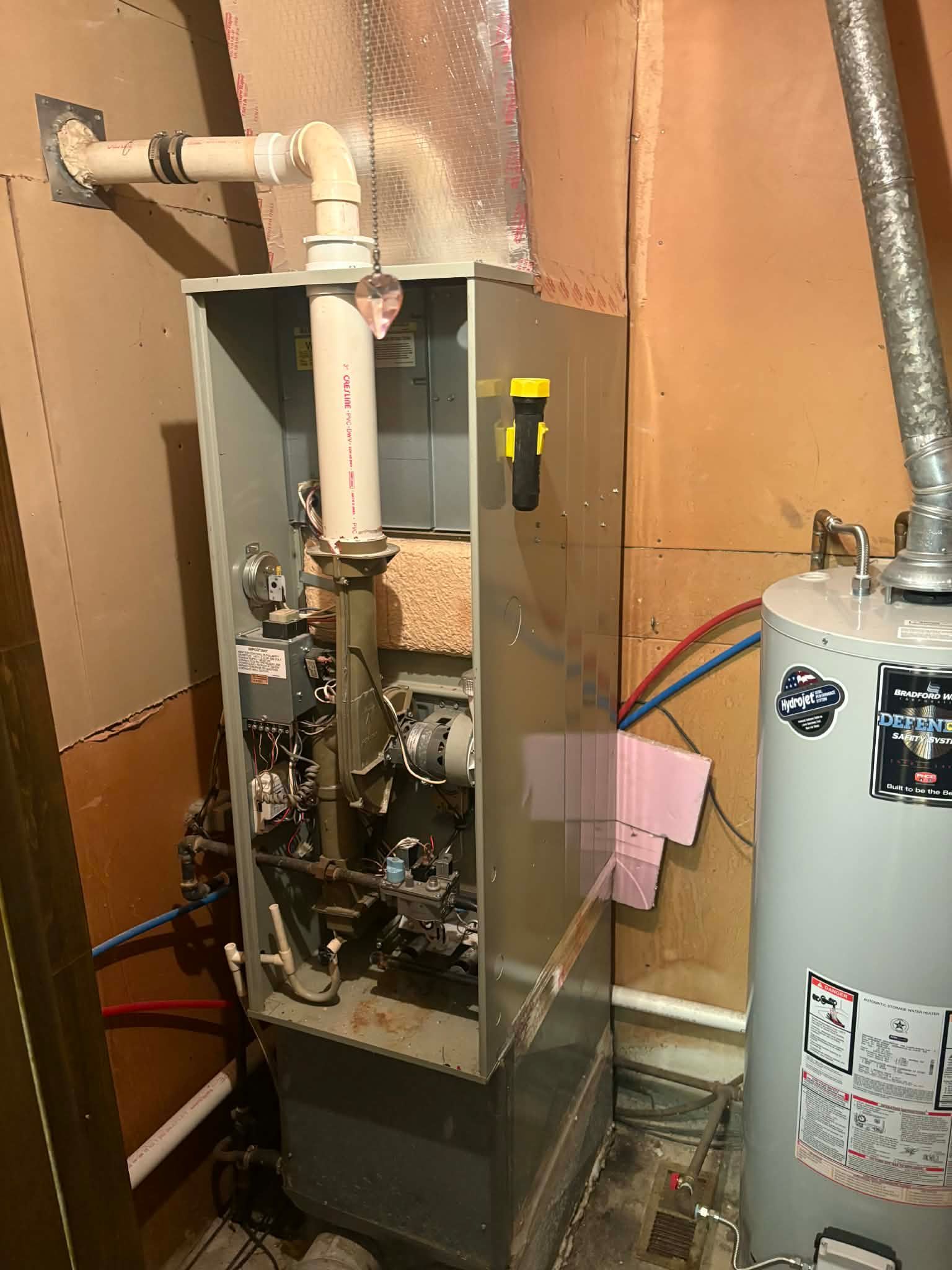 A grey vertical gas furnace with a white PVC vent pipe stands next to a cylindrical water heater in a utility closet.